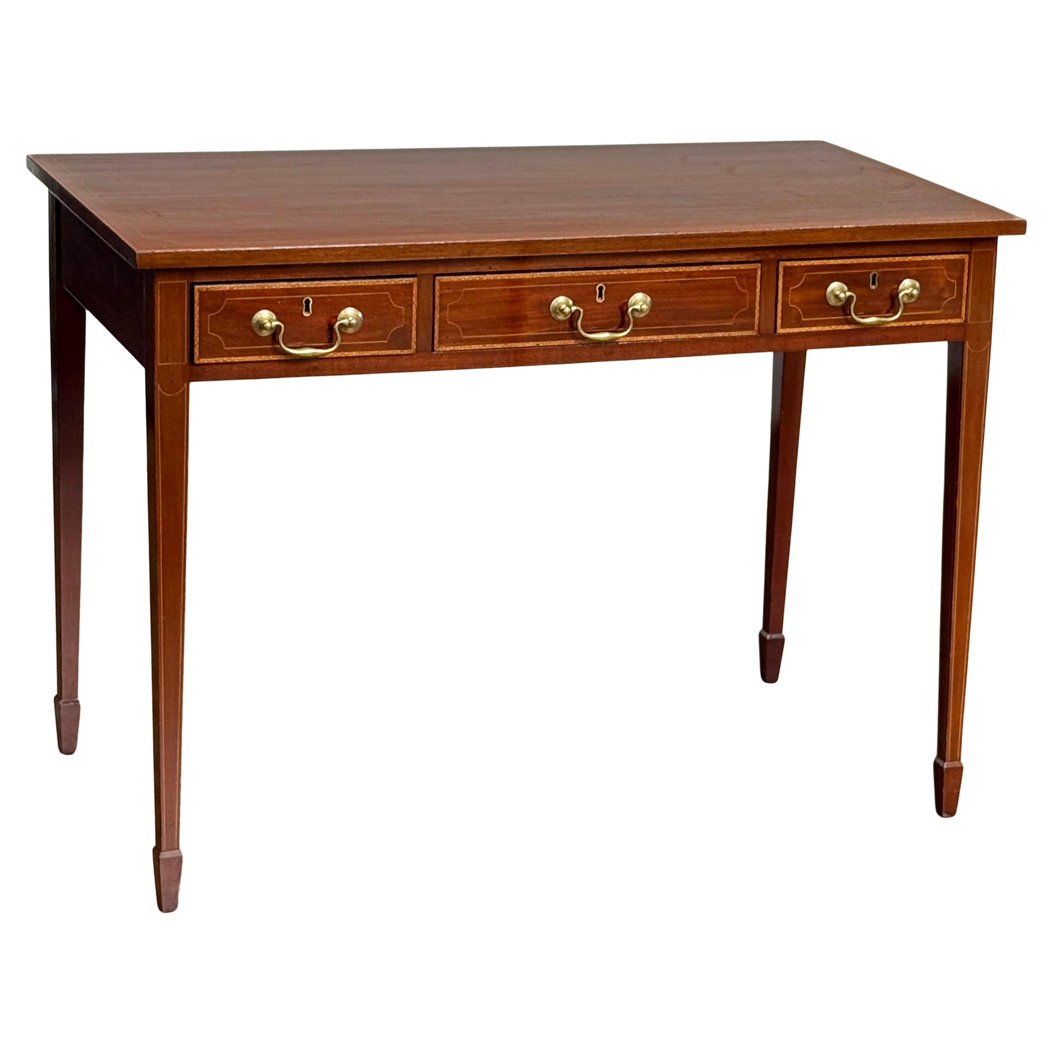 A fine English writing table or desk of mahogany from the Edwardian Era, featuring a rectangular top with inlay along the front edge, over a frieze of three drawers - each with brass swan neck handles, with decorative inlay to drawer facings and to
