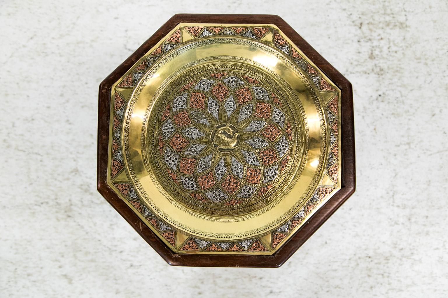 This engraved brass tray table has an engraved octagonal brass tray that is overlaid with raised copper and silvery metal panels that are engraved and chased with arabesques. The center of the tray has a raised stylized engraved bird. The solid