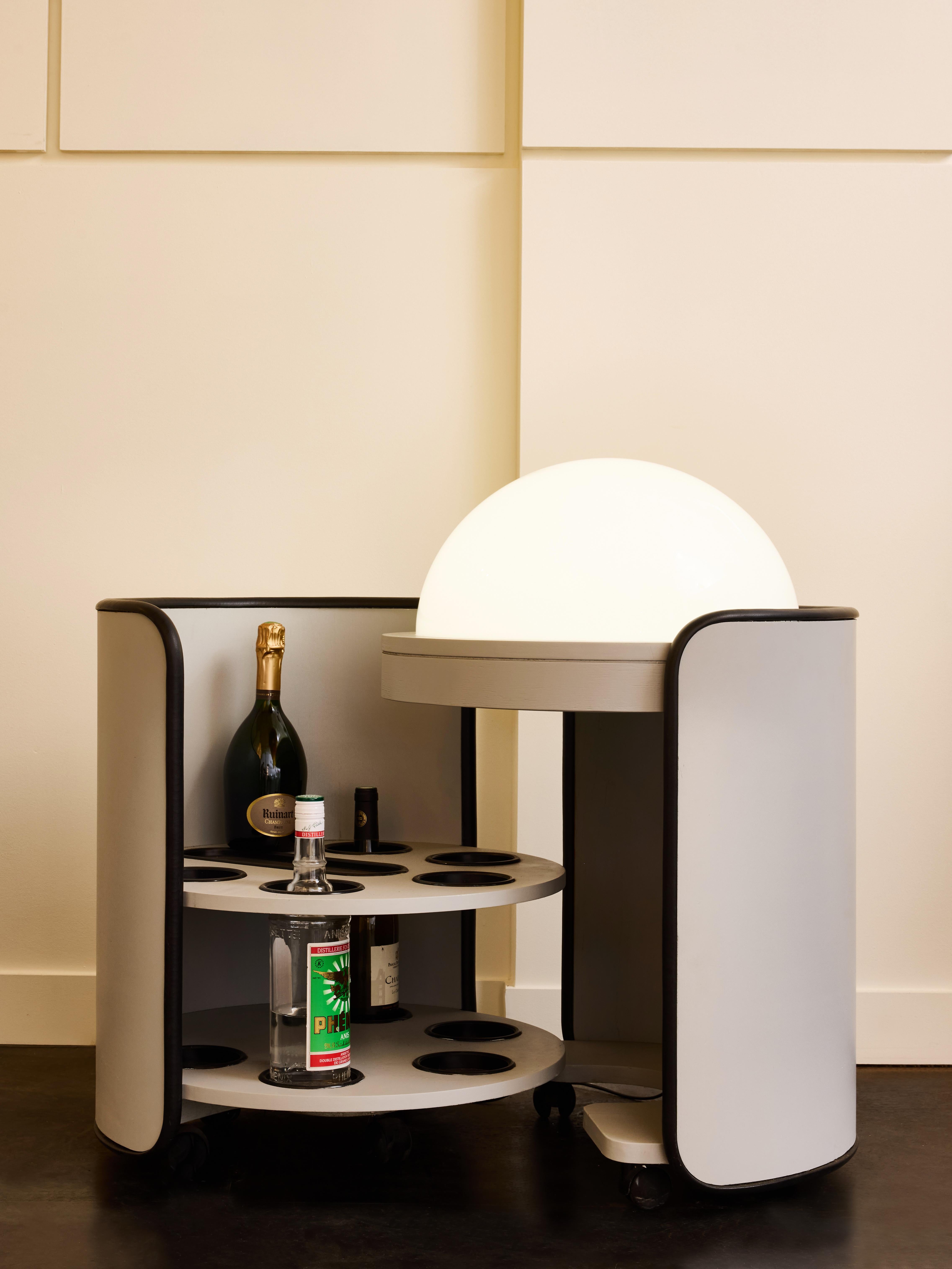Una lampada da terra rotonda in legno sormontata da una semisfera in plexiglass completa di interruttori originali. La lampada si apre senza sforzo grazie alle ruote integrate, svelando un bancone asciutto che ospita cinque bottiglie e bicchieri,
