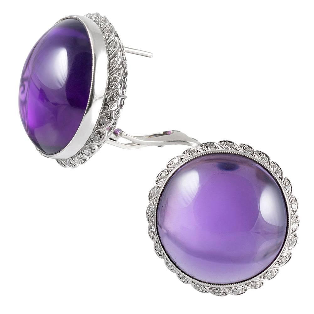 These striking earrings are set in the center with extra-large cabochons of amethyst that measure 19 millimeters in diameter. The handmade platinum frames boast an artful scalloped border with diamonds surrounded by millgrain edges. In total, there