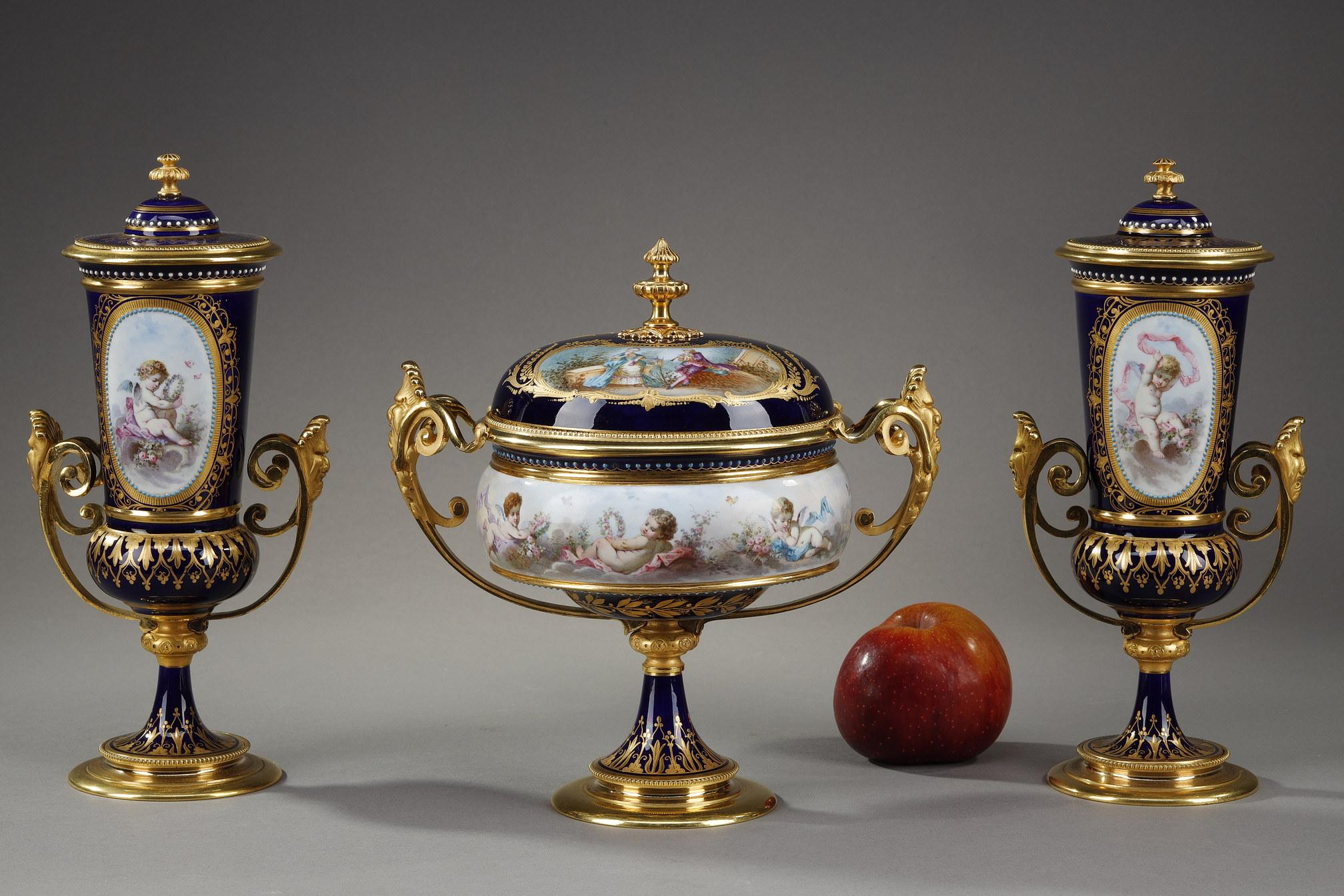 Ensemble aus einem Becher und zwei Vasen aus Sevres-Porzellan mit blauem Hintergrund. Die Elemente sind in vergoldeter und ziselierter Bronze gefasst, deren Griffe mit grotesken Masken und Schriftrollen verziert sind. Der Hintergrund ist mit