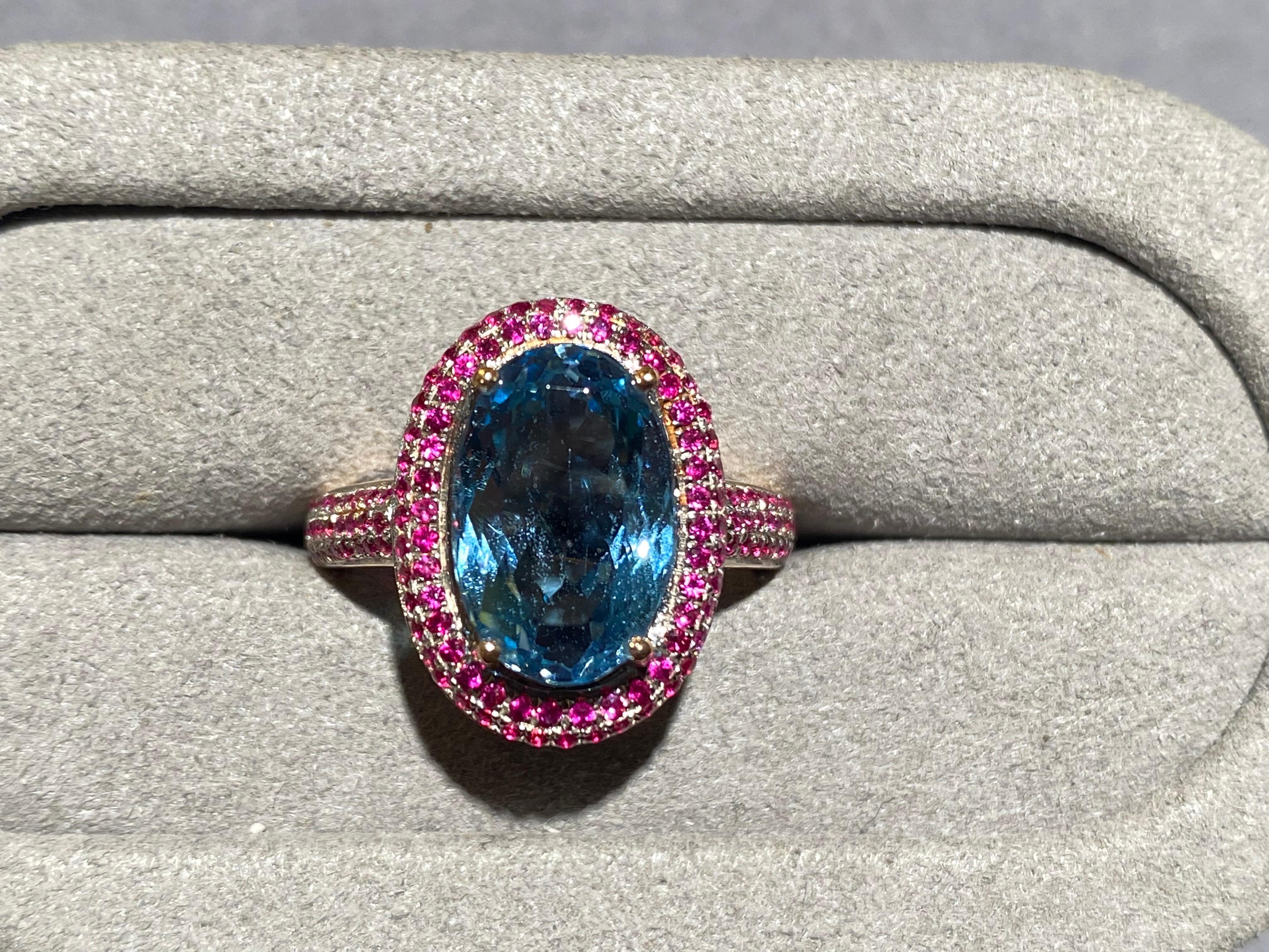A Vivid blue aquamarine and ruby ring in 18k rose gold. It is an oval shape cut aquamarine surrounded by micro ruby pave. Half of the ring band is also set with micro rubies. This is a very bold choice of colour as vivid blue and vivid red they