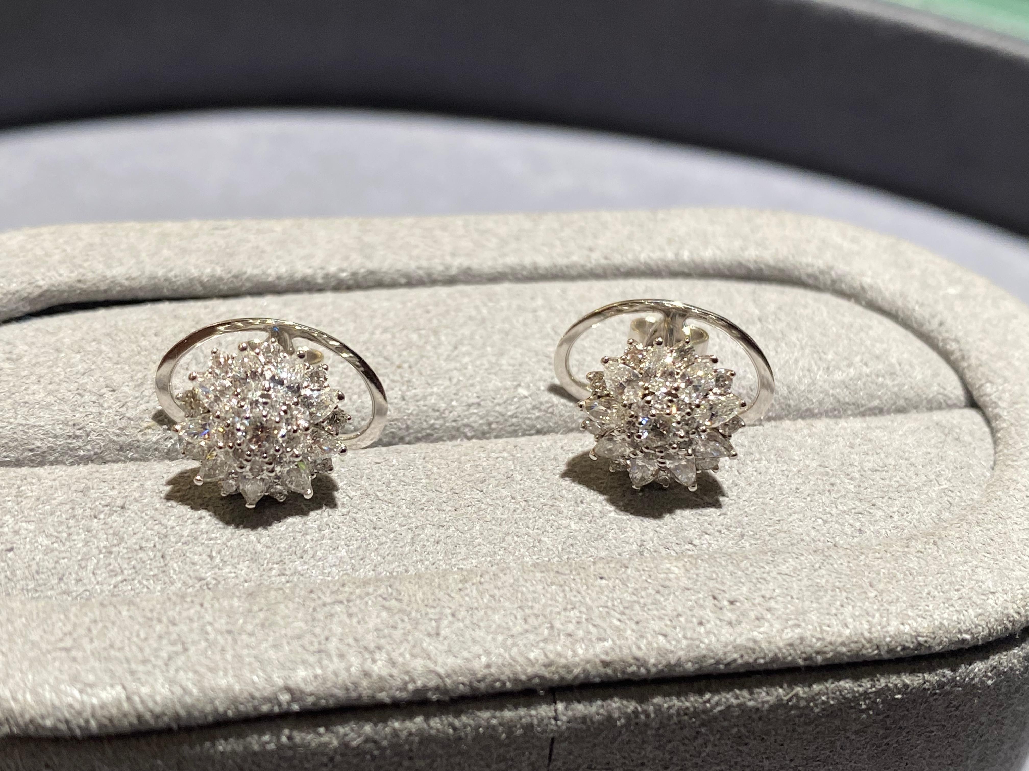 A pair of flower motif diamond earrings in 18k white gold. A round brilliant cut diamond is set in the middle of the flower motif with a circle of micro diamonds surrounding the middle. On the outer layer there are multiple rain drop diamonds and in