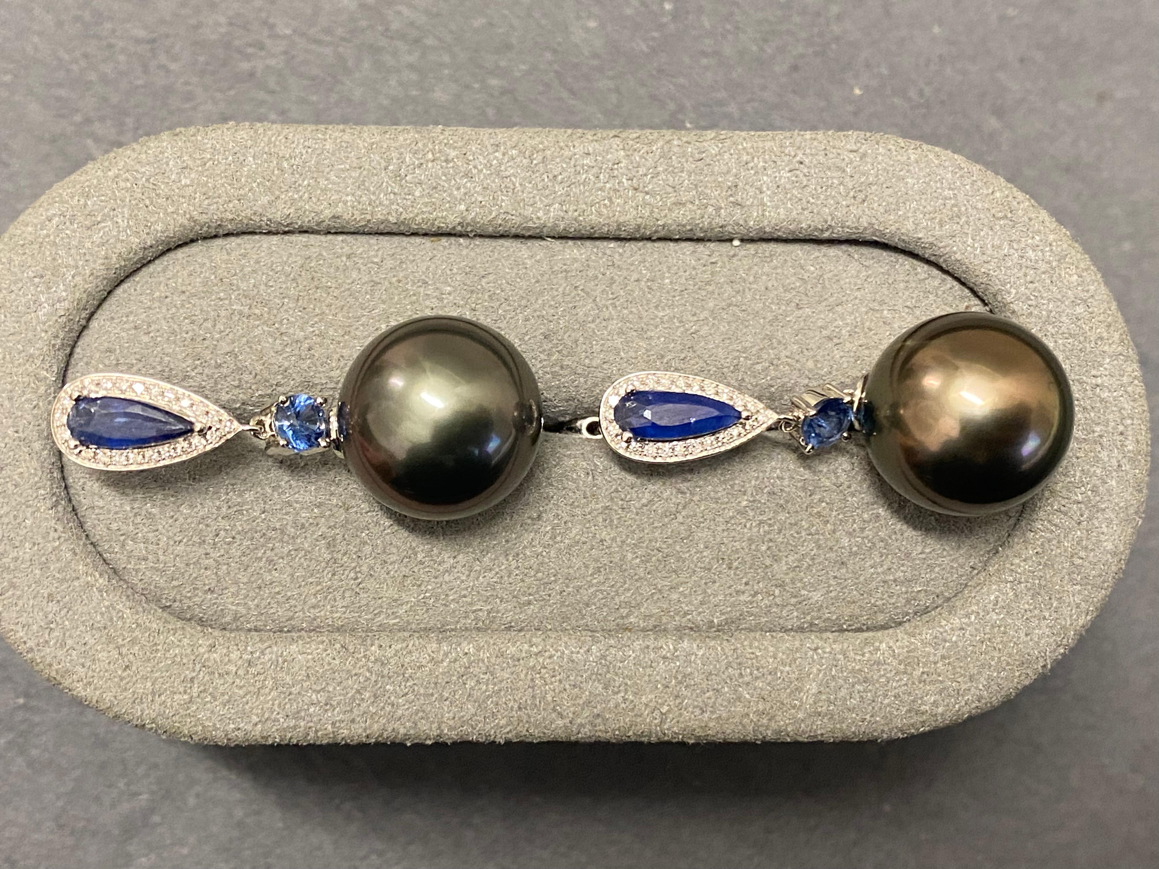 A pair of Tahitian pearl, blue sapphire and diamond earrings in 18k white gold. The Tahitian pearl is suspended below a rain drop shape blue sapphire and is connected via a round lighter colour blue sapphire. This set up allows the dangle Tahitian