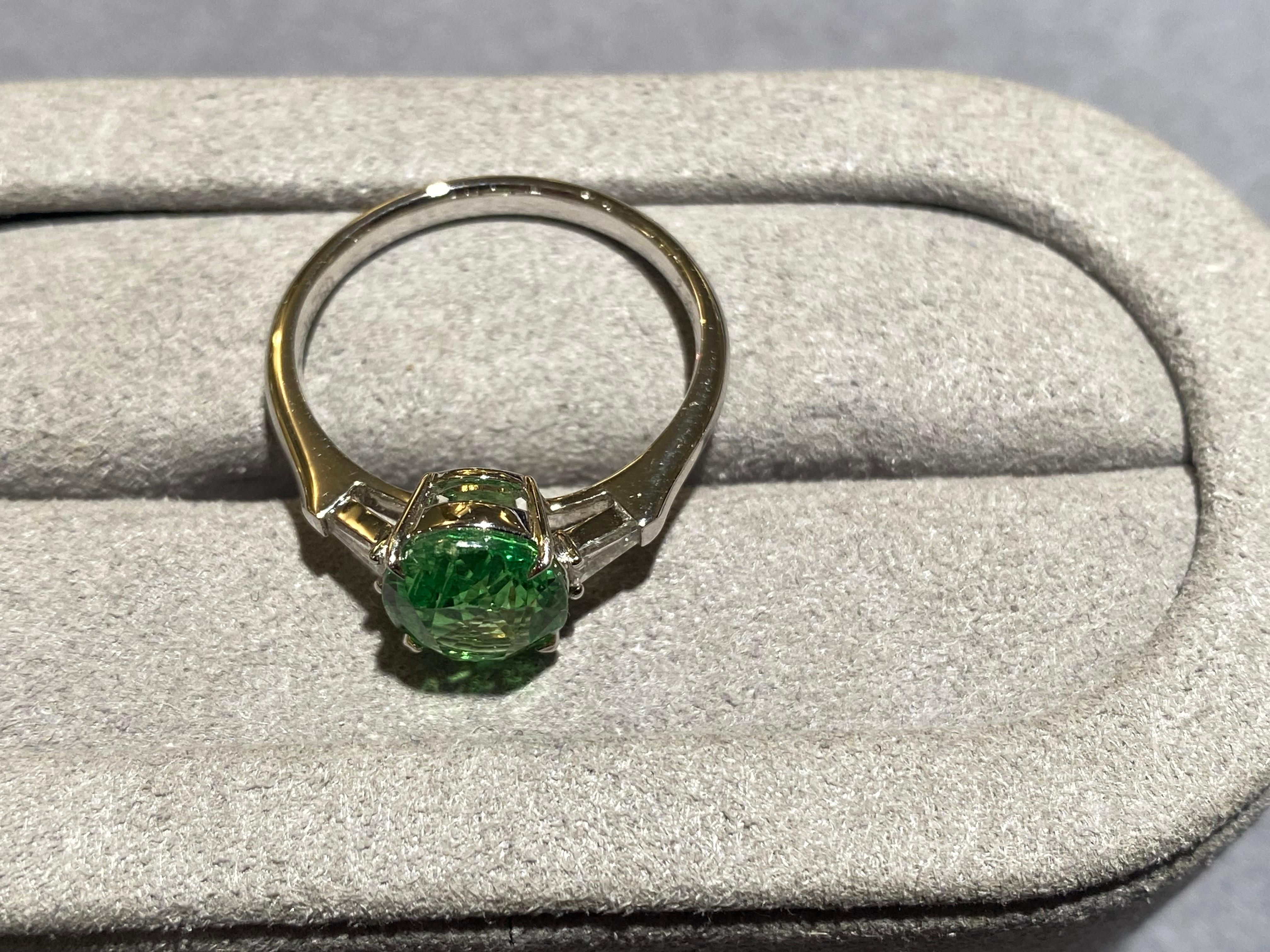 A Tsavorite and diamond three-stone ring in 18k white gold. The main tsavorite is accompanied by 2 tapered baguette diamonds. The Tsavorite is round in shape and with pastel green colour. This is a very simple yet elegant design perfect for everyday