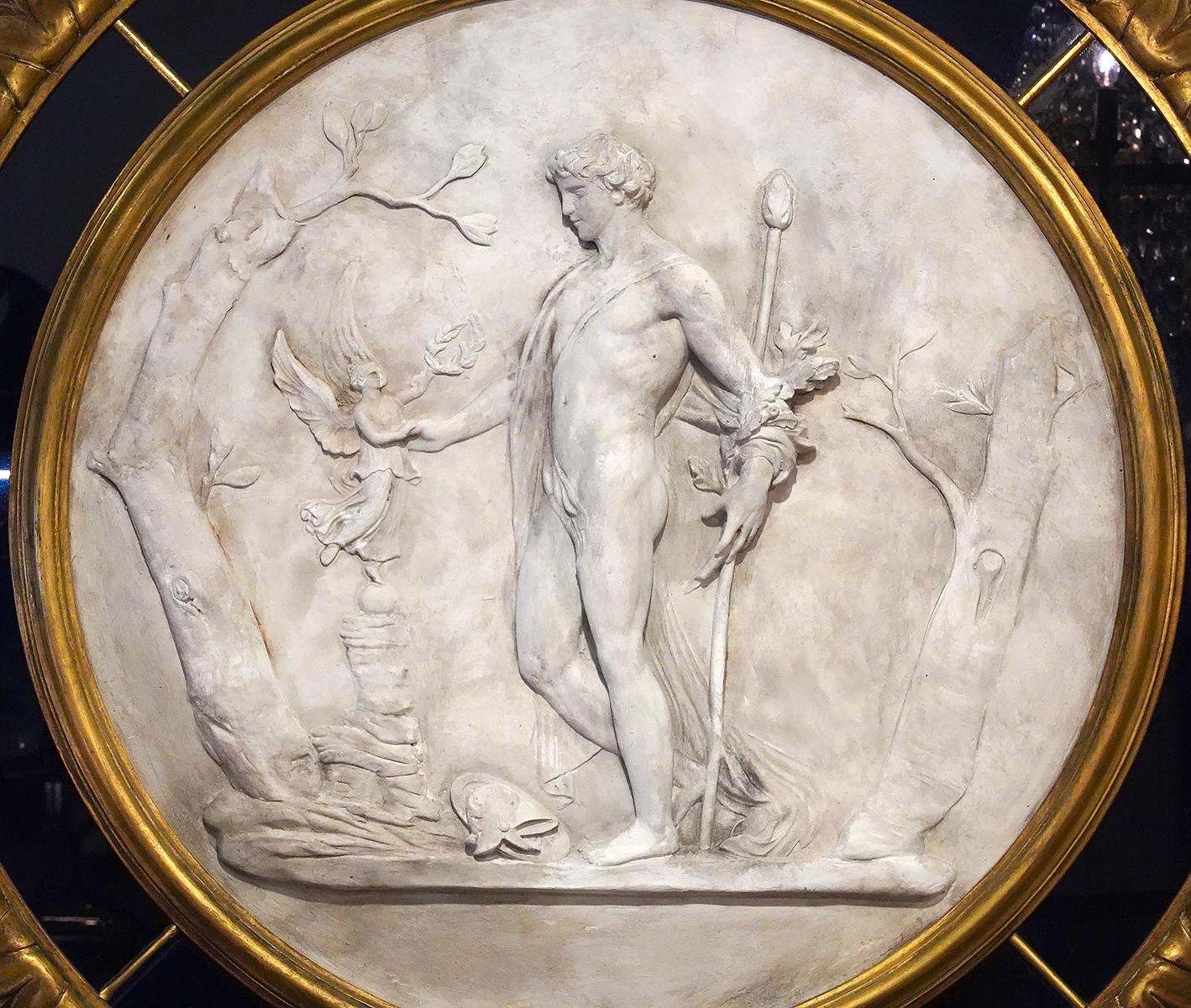 This Italian classical plaster relief likely after a marble original depicts a Roman hero, his shield and helmet on the ground, being offered a laurel wreath from an angel standing on a small sphere. The motif is flanked by rudimentary trees. The