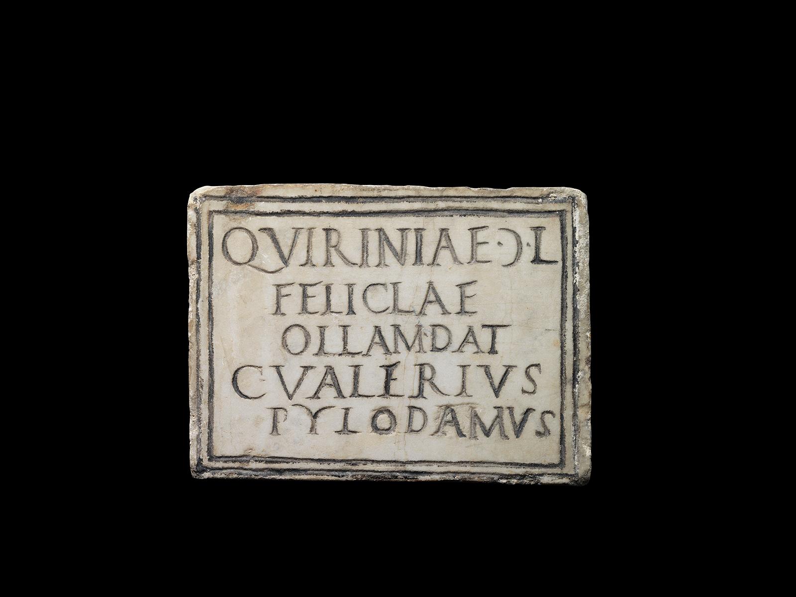 A rectangular marble slab carved with the Latin inscription ‘QVIRINIAE C(retr.) L / FELICLAE / OLLAM DAT / C VALERIVS PYLODAMVS’, which translates as ‘Gaius Valerius Pylodamus gave the burial urn to the freedwoman Quirinia Felicia’. This is a tablet