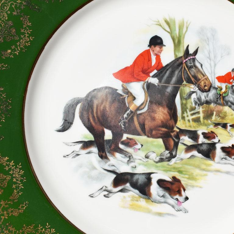 A green and gold ceramic plate of an equestrian hunting scene. This lovely piece features a thick green border with gold detail. The center of the plate depicts a team of hunters on horseback in red coats amidst hunting dogs. 

Measures: 10.5