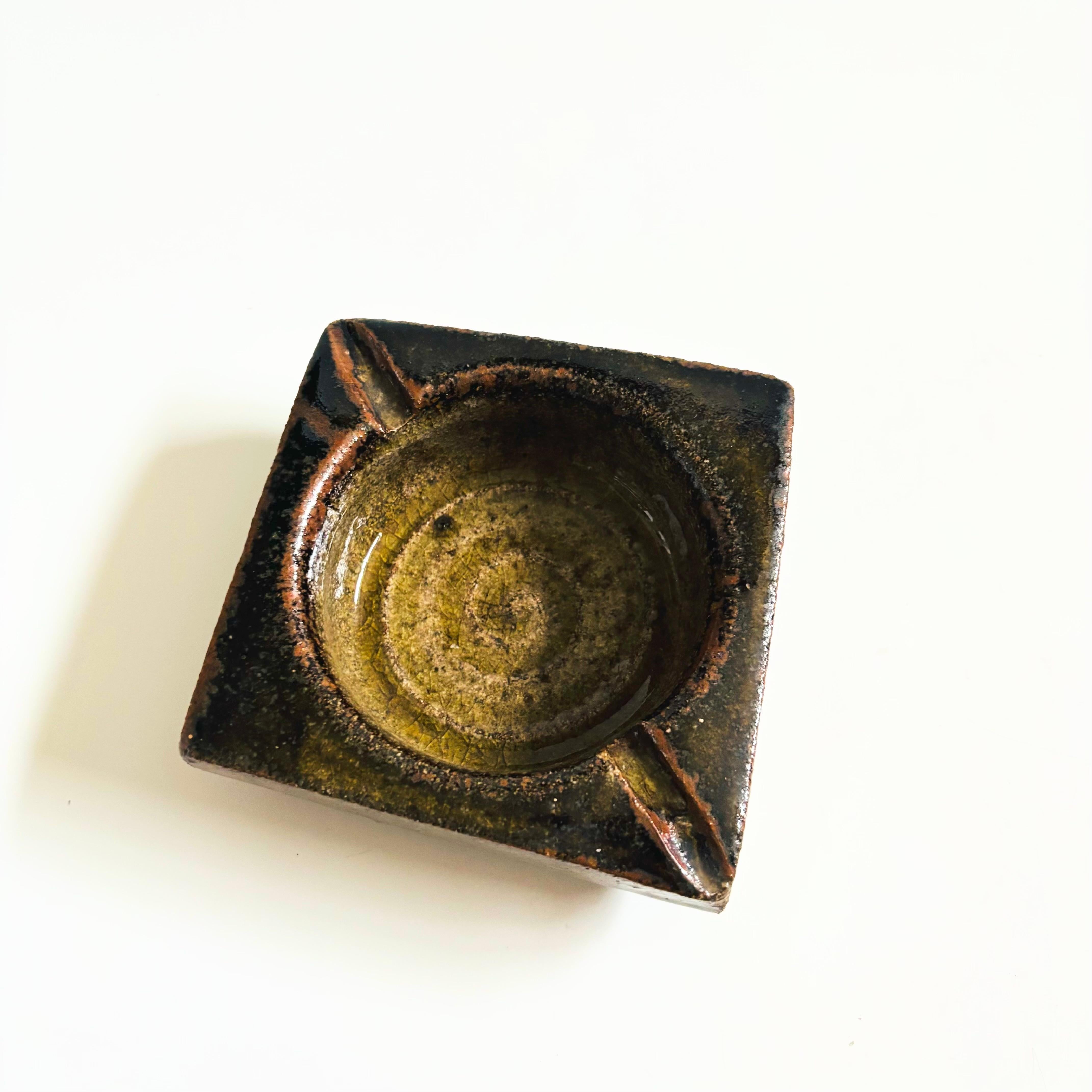 Cendrier vintage en poterie de studio du céramiste californien Eric Norstad (1924-2013). Forme carrée irrégulière avec un bol circulaire et deux fentes pour cigarettes. Finition en glaçures de couleur terre. Estampillé sur la base 