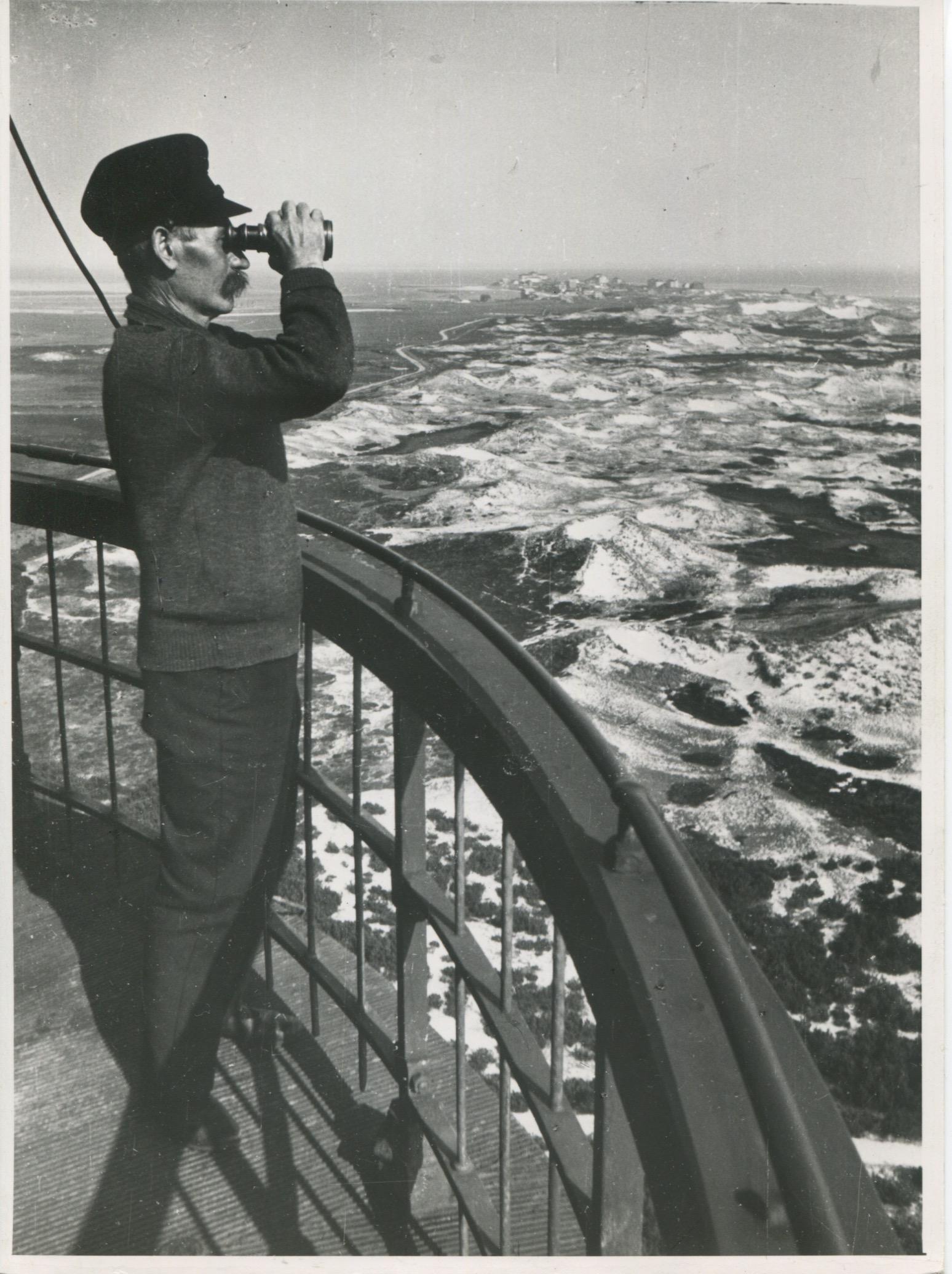 Grabado en gelatina de plata de Erich Andres, 1941.

Esta fotografía, tomada en 1938 en Amrum, muestra a un farero de pie en la plataforma de observación del faro. Lleva uniforme y gorra, típicos de la época, y contempla el paisaje costero a través