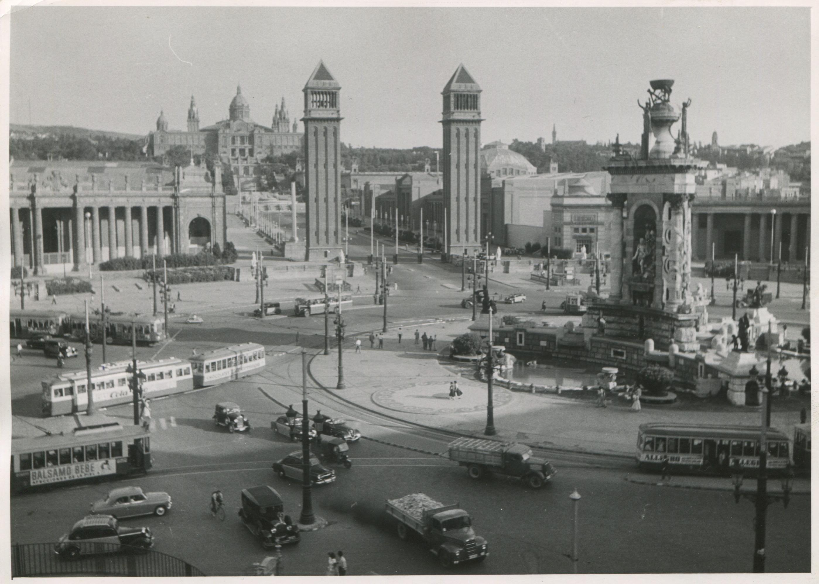Silver Gelatine Print by Erich Andres, 1957.

This black-and-white photograph from 1957 captures the bustling Plaza de España in Barcelona, Spain. In the foreground, a grand traffic circle surrounds a large fountain adorned with sculptures and