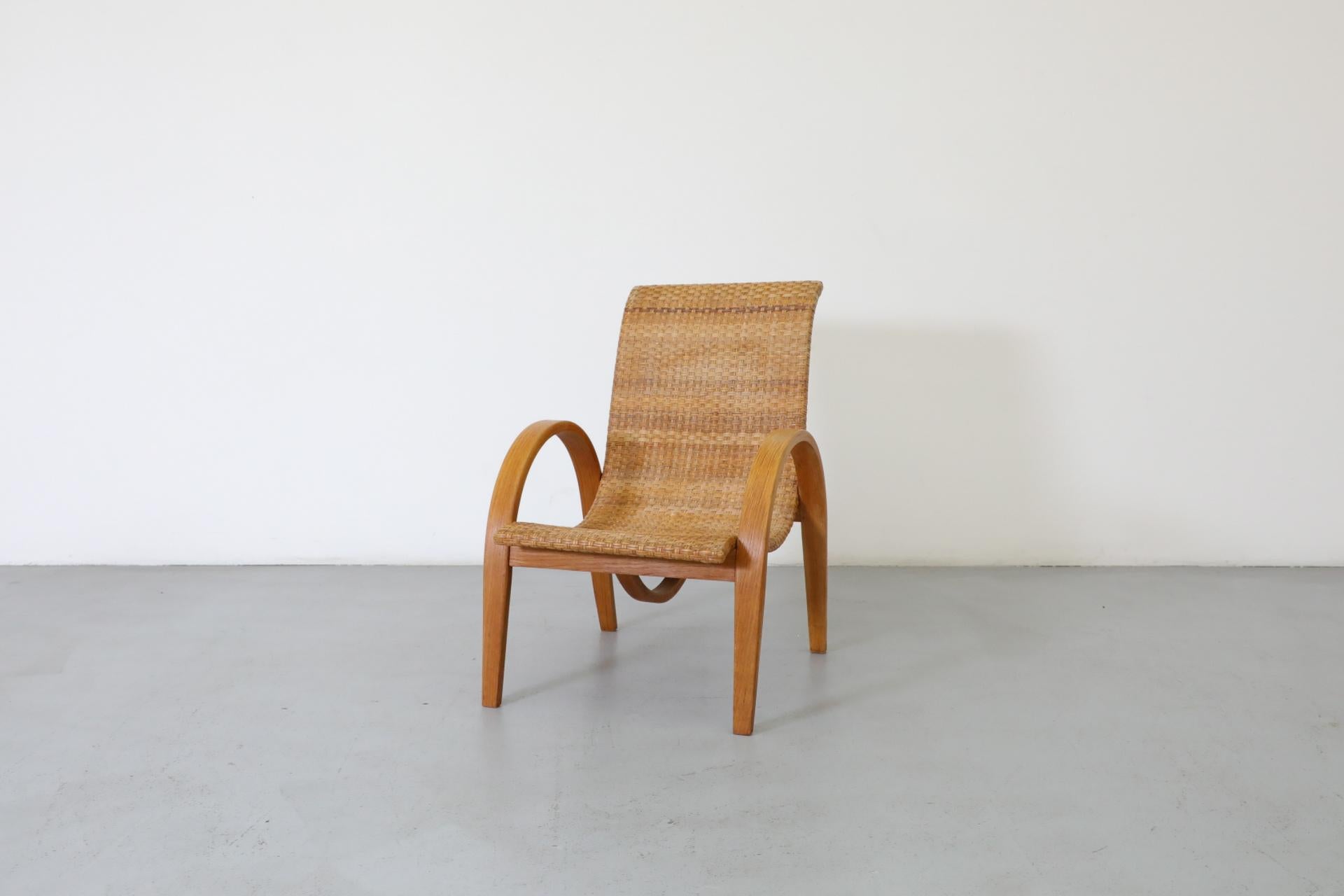 Tumbona de ratán tejido, estilo Erich Dieckmann, con gruesos reposabrazos de roble bellamente curvados y soportes curvados a la vista. Elegante y cómoda, la silla también es muy similar a algunos ejemplos modernistas tempranos de sillas de madera y