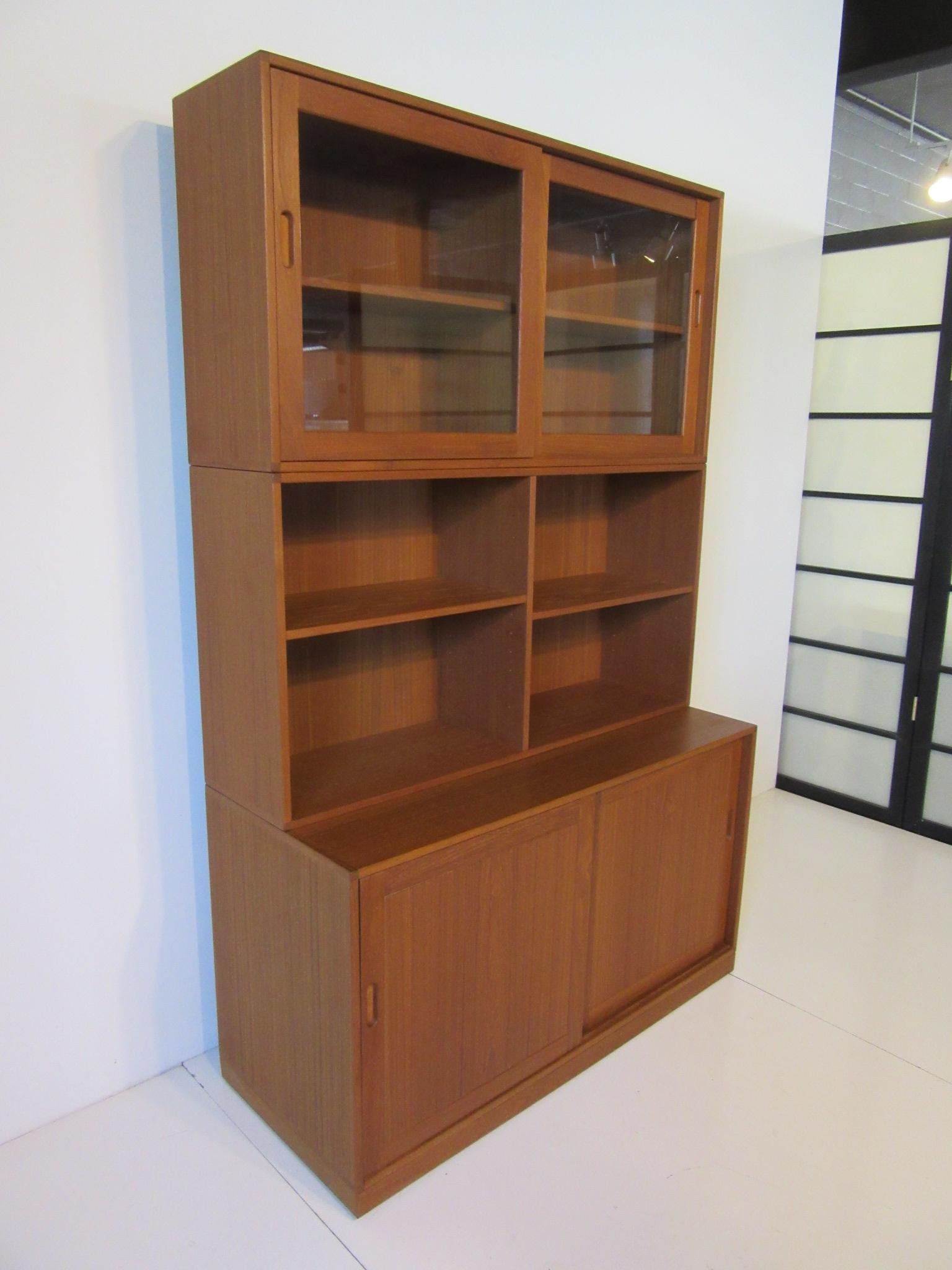 A well constructed solid teak wood bookcase / cabinet with upper sliding glass doors, adjustable shelves with lower sliding doors and storage, shelves including a small drawer. The inset door pulls are leather lined showing the quality and