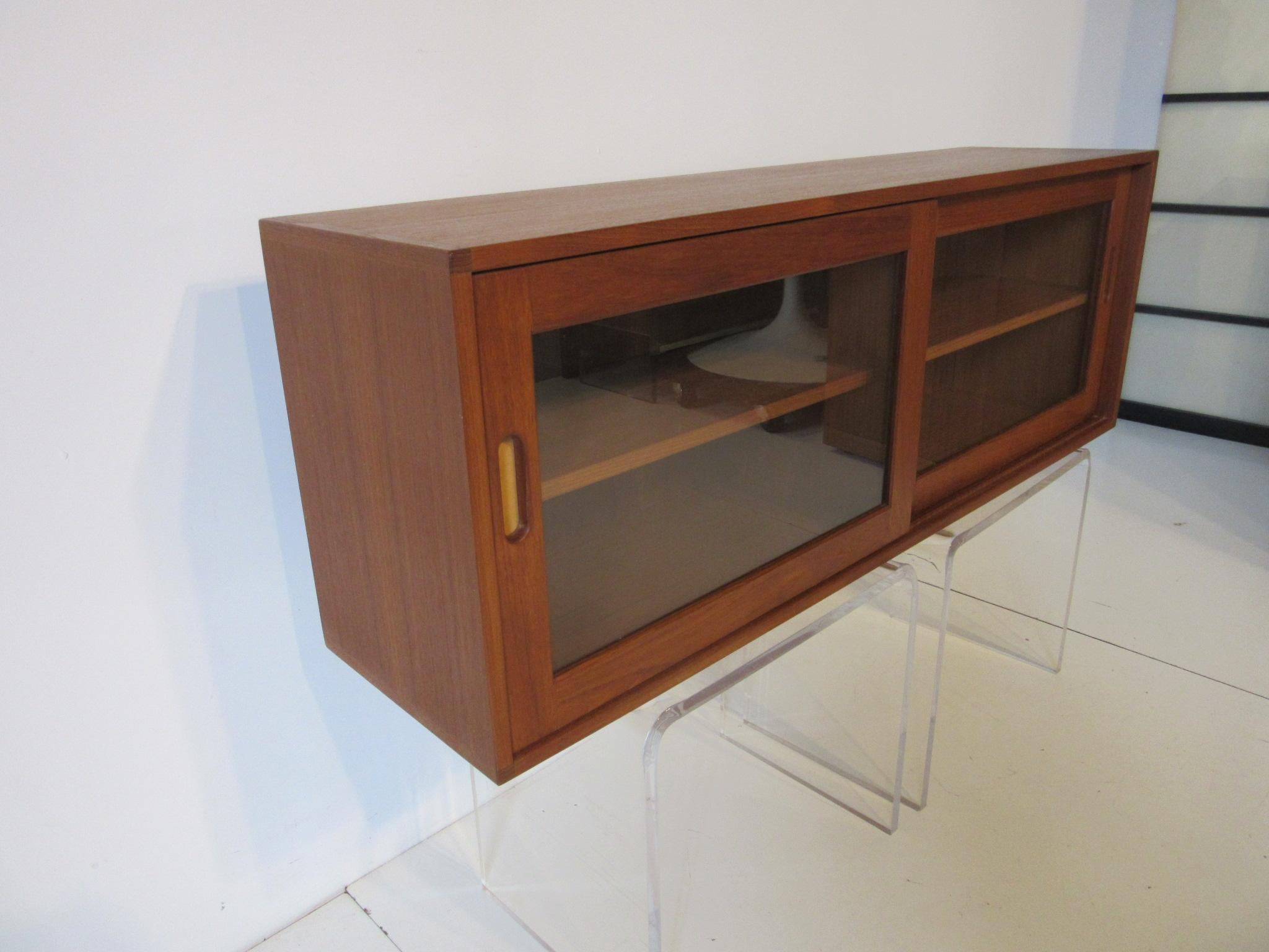 A well crafted two sliding door teak and glass hanging cabinet with two adjustable shelves, leather inserts to the wood craved pulls made in Denmark by Brouer Mobelfabrik. Purchased in Denmark in 1962 by the original owners and lovingly cared for