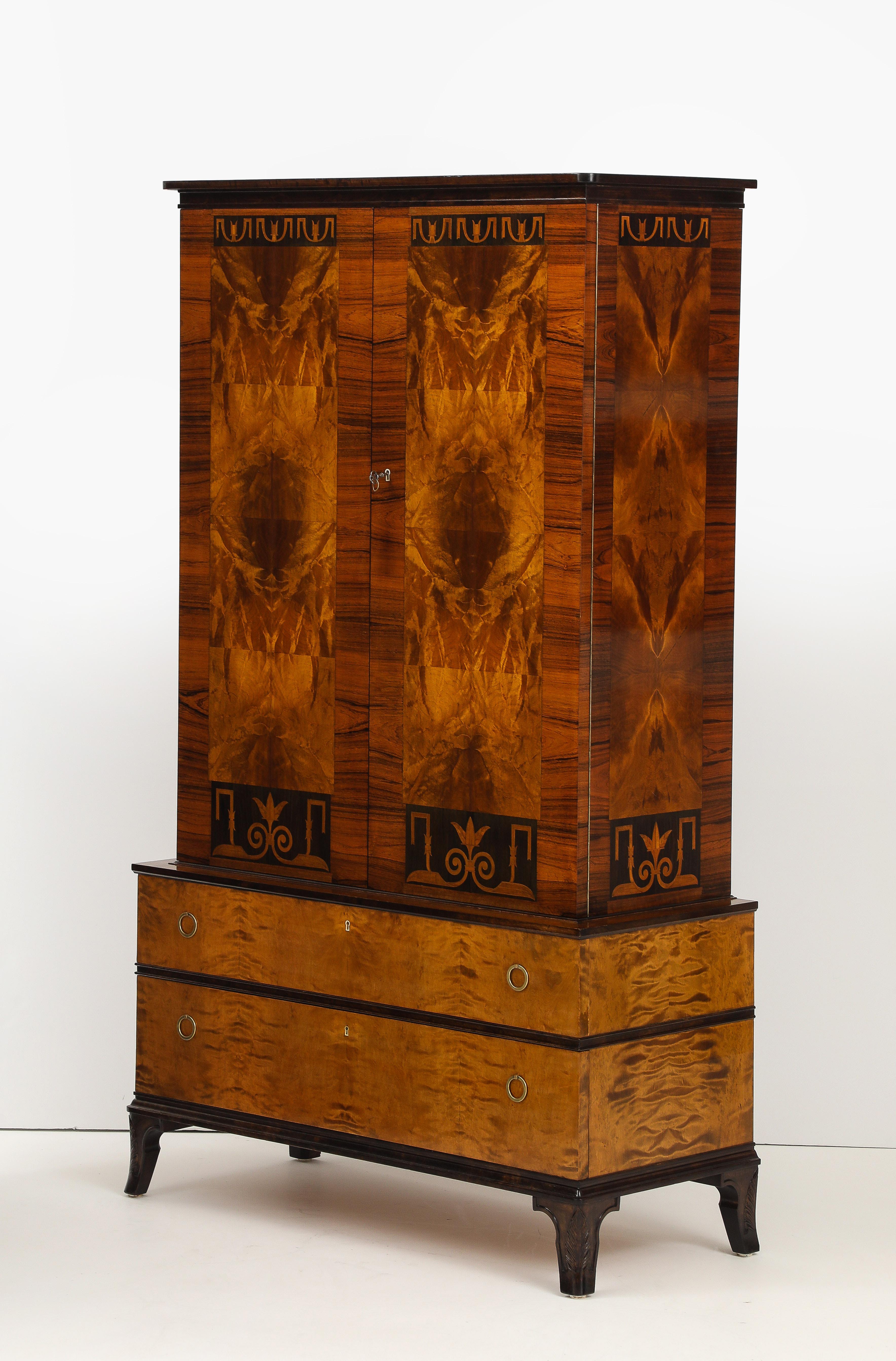 A Swedish birchwood, walnut and palisander cabinet by Erik Chambert, Circa 1920s, produced by Chamberts Möbelfabrik , the upper half with an overhanging cornice molding above two cupboard doors with Swedish Deco inlay and vibrant book matched