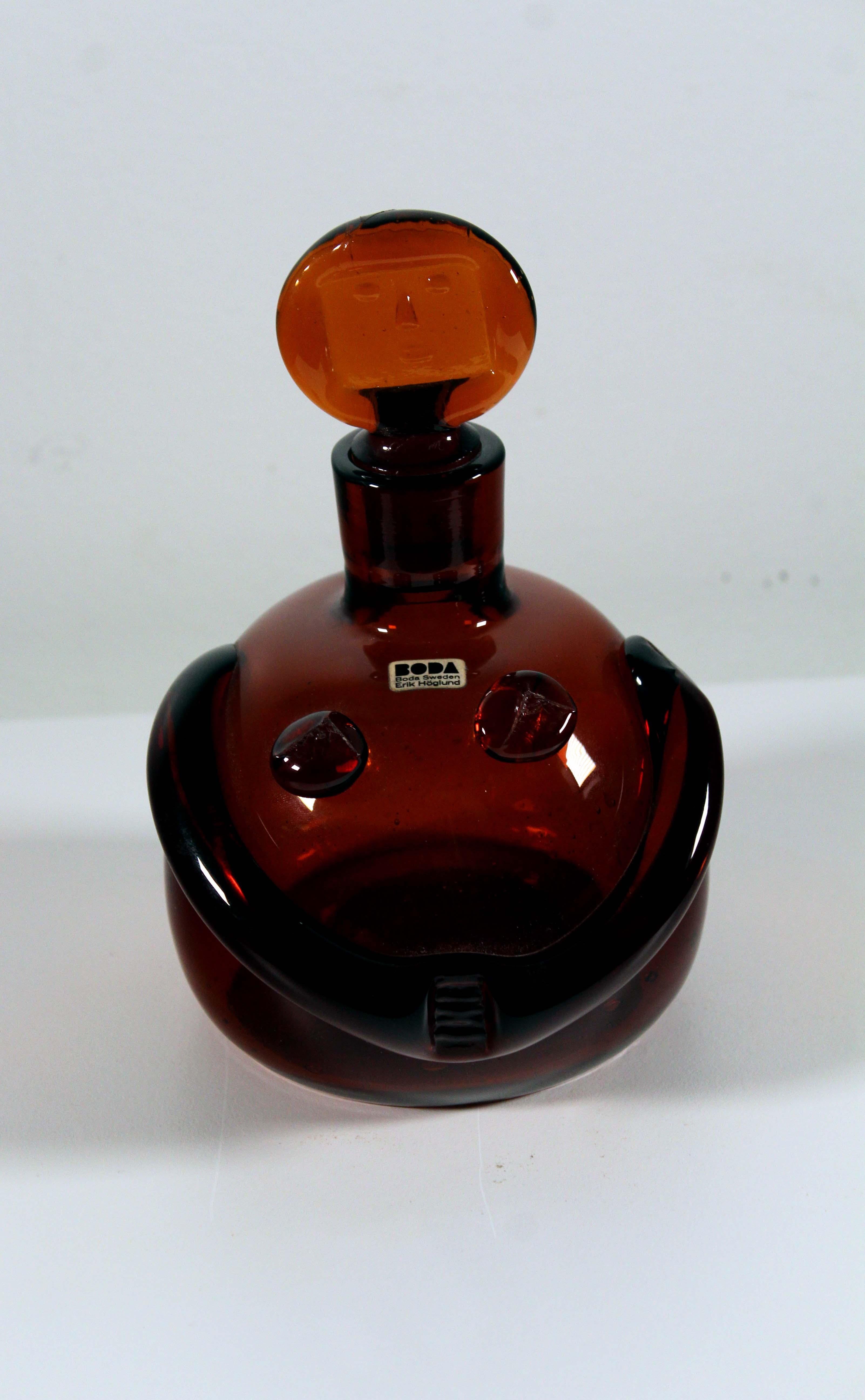 An iconic Mid-Century Modern orange glass decanter by Swedish artist Erik Hoglund. The lid of the bottle has his infamous female face design. With original Boda Sweden product tag. From a private collection. Dimensions: 6.5” height x 4.5” diameter