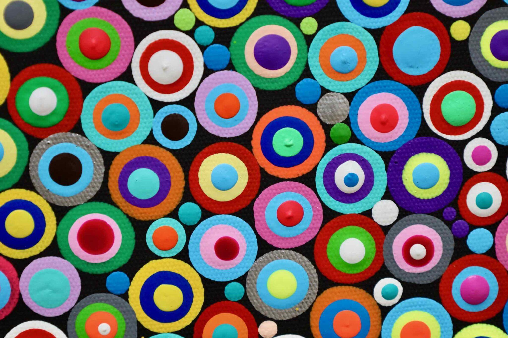 “Corollary” by Ernesto Guevara features a large, perfectly round circle composed of countless small, vibrant circles on a deep black background. Each small circle contains various colors, creating a mosaic of dots that almost shimmer like colored