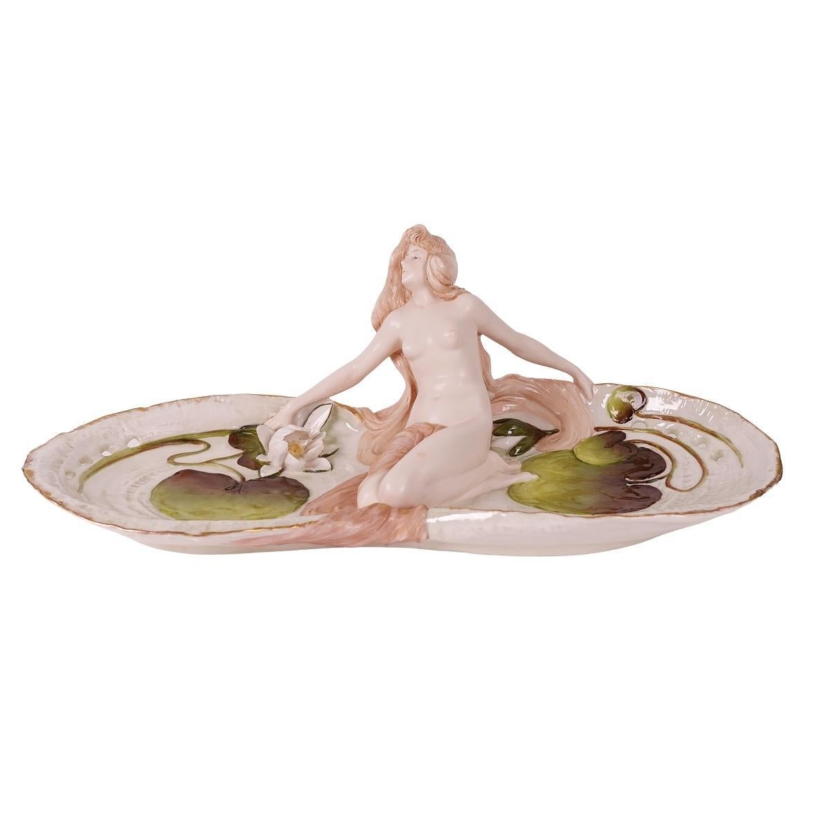 Nous vous proposons ce magnifique plateau figuratif en porcelaine peint à la main par Ernst Wahliss, de style Art nouveau. Ce plateau représente une jeune fille nue assise, flanquée de motifs aquatiques en forme de nénuphars verts et d'une fleur