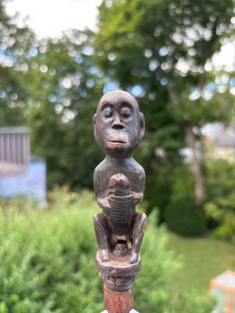 Sculpture érotique indonésienne représentant un homme tenant son phallus surdimensionné en érection, assis en position accroupie. La sculpture est le manche d'un outil en fer, probablement une herminette pour la sculpture. L'expression sérieuse du