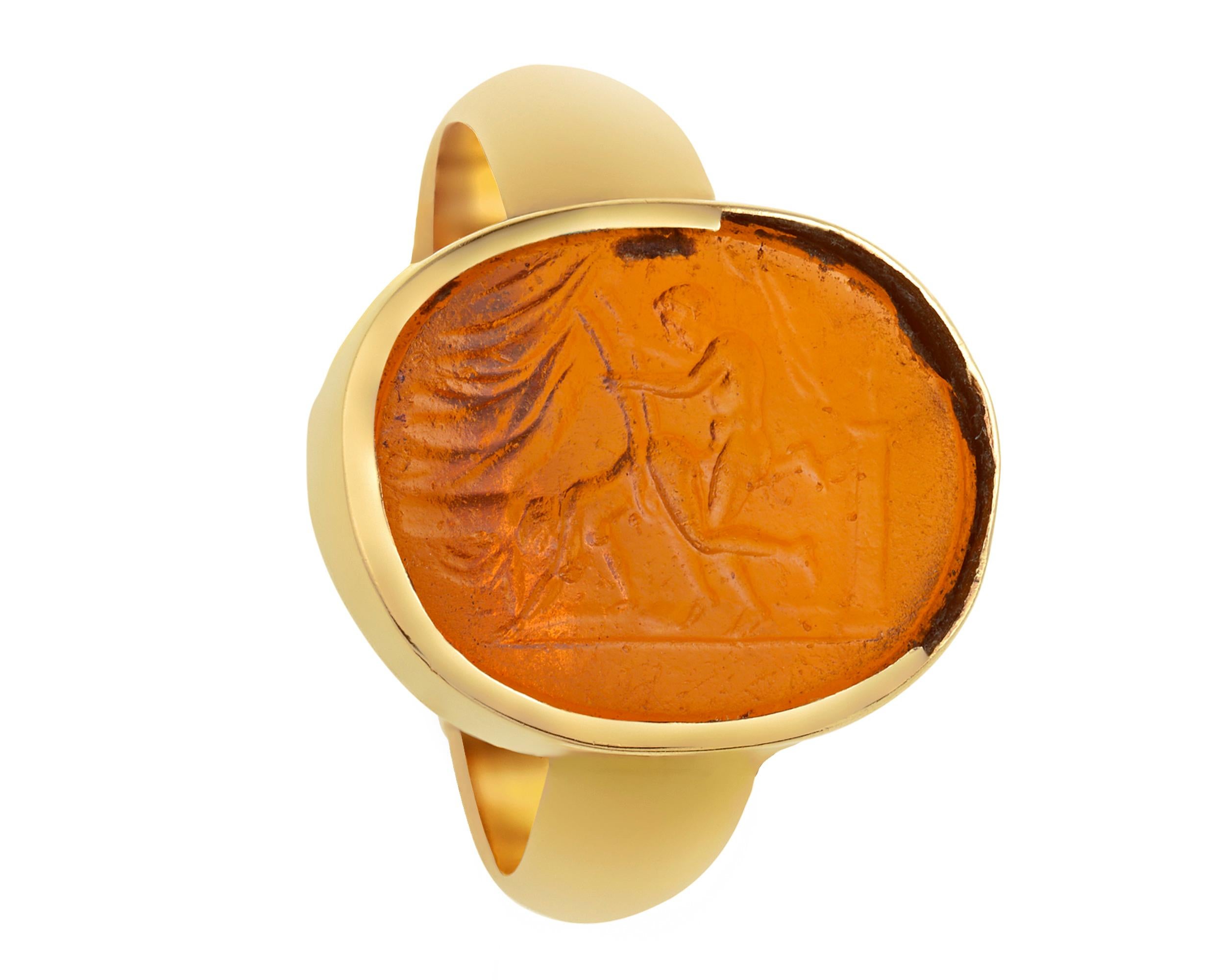 Beautifully formed and enticingly erotic, this glass ring begs closer inspection. Vibrant orange glass in a 14K yellow gold oval-shaped bezel setting provides a visually striking first impression. A closer look, however, reveals an intaglio image of