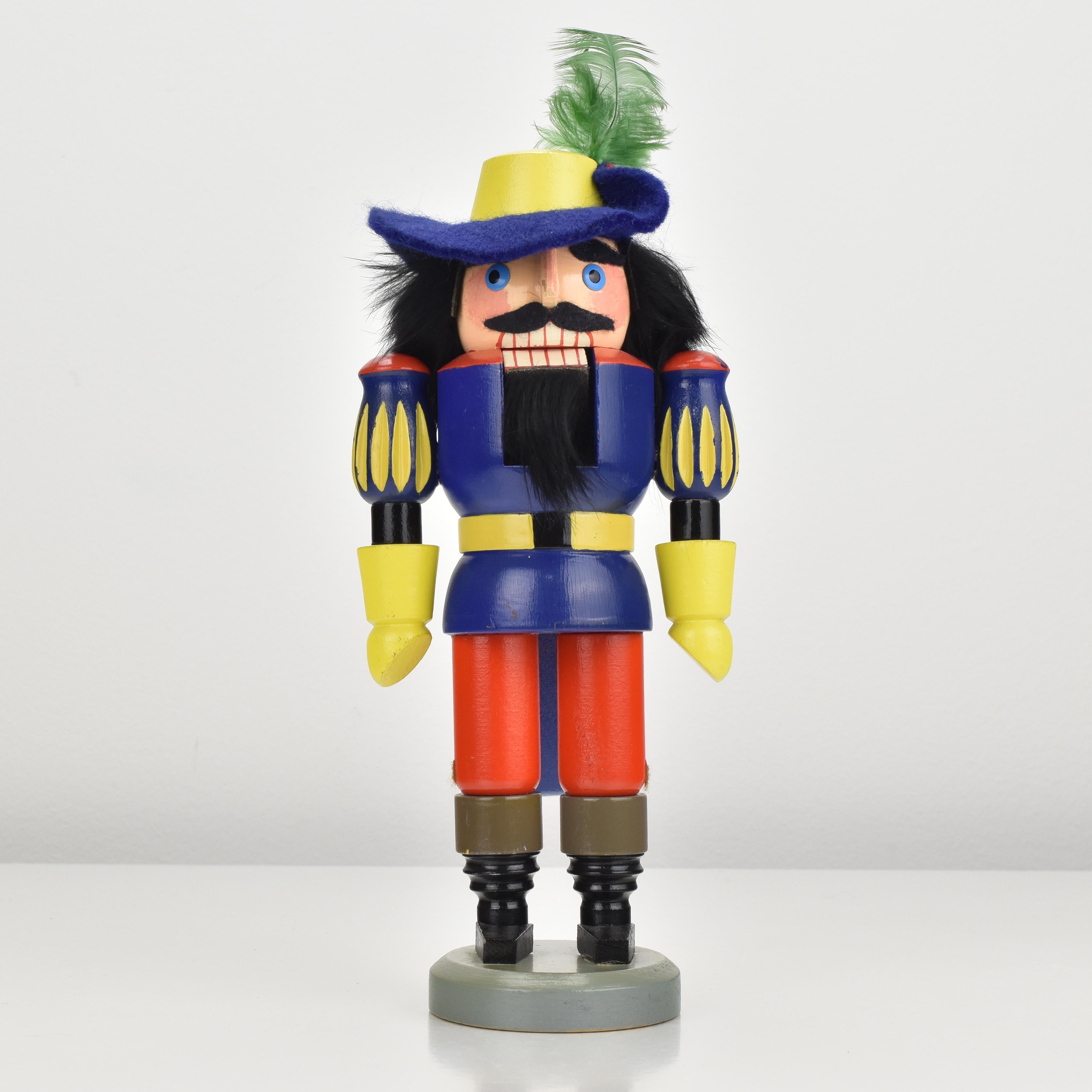A vintage nutcracker from the Erzgebirge region, in shape of a robber or nobleman made of turned wood and painted in vibrant colors, dating back to the 1970s.
This nutcracker features a blue felt hat with a green feather and is decorated with real