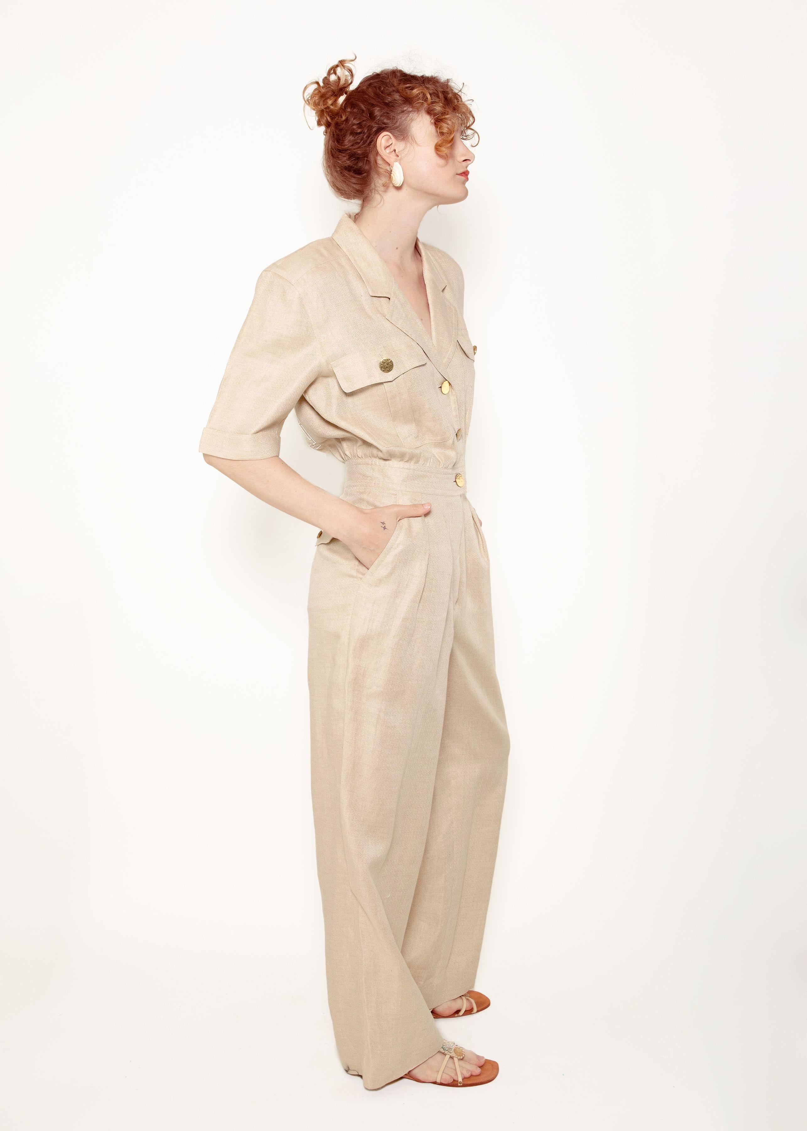This Escada Linen Jumpsuit is perfect for a summer day. Made from lightweight sand colored linen, the jumpsuit features brass hammered buttons, front and side pockets, short sleeves, and a v neck with a collar. Stay cool and comfortable all season