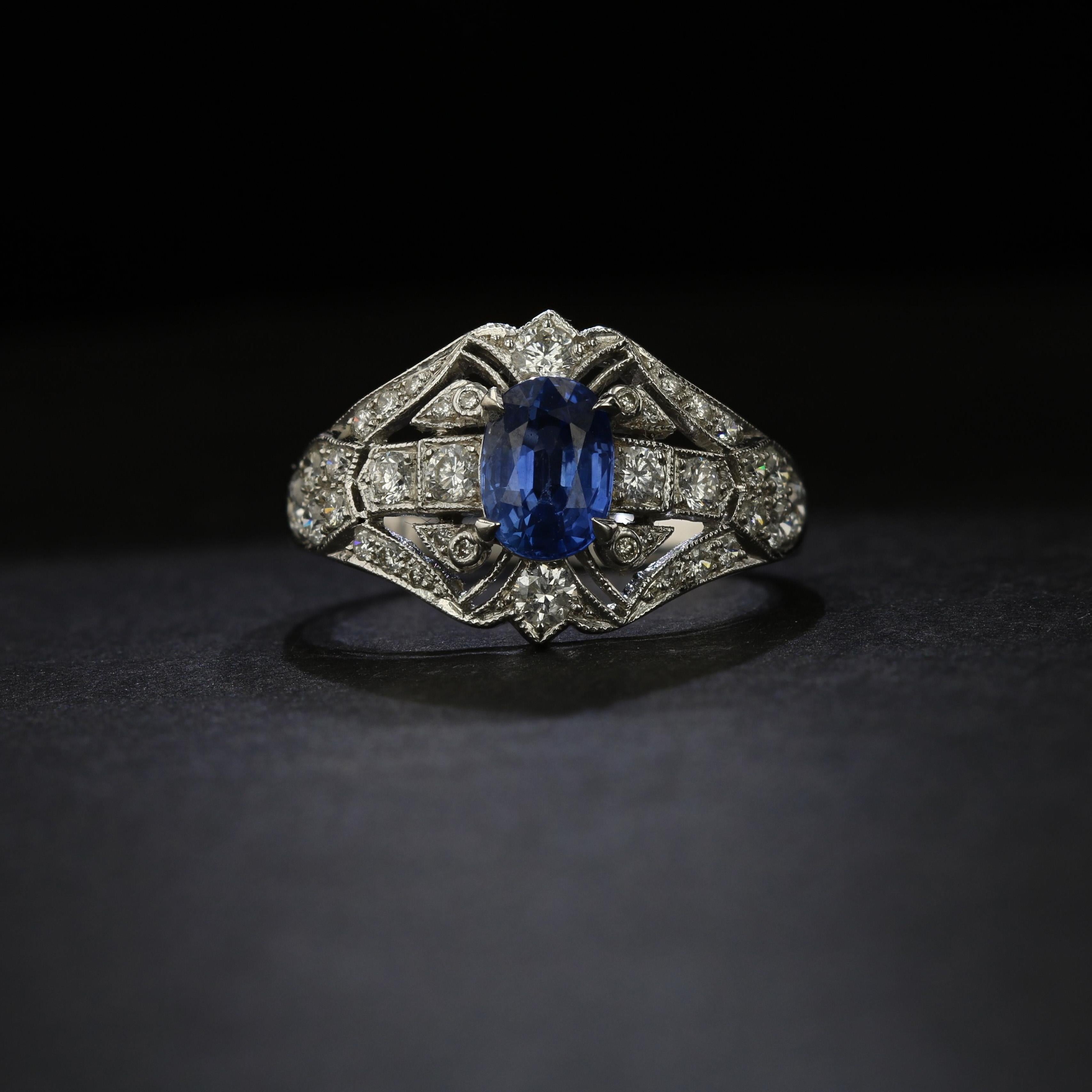 This 14 K white gold Art Deco style ring contains one 1.35 carat oval blue sapphire set by 4 prongs in the center.  The ring features millgrain and filigree details, and is set with an additional 1.35 carats of round diamonds.  The diamonds have an