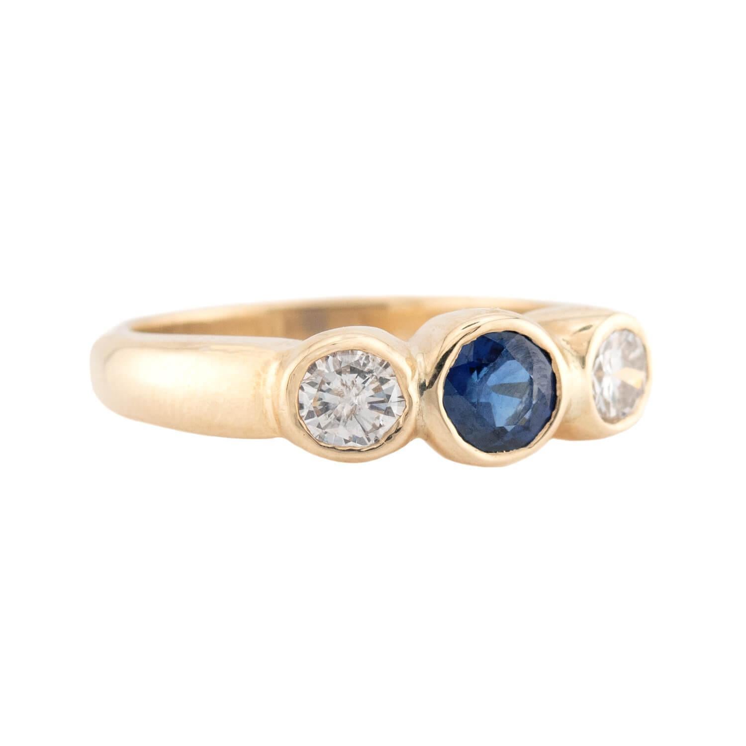 A beautiful sapphire and diamond ring! This gorgeous piece is crafted in 14k yellow gold, and features three stones running horizontally across the front of the ring. The center stone is an round cut sapphire, which is held within a bezel setting.