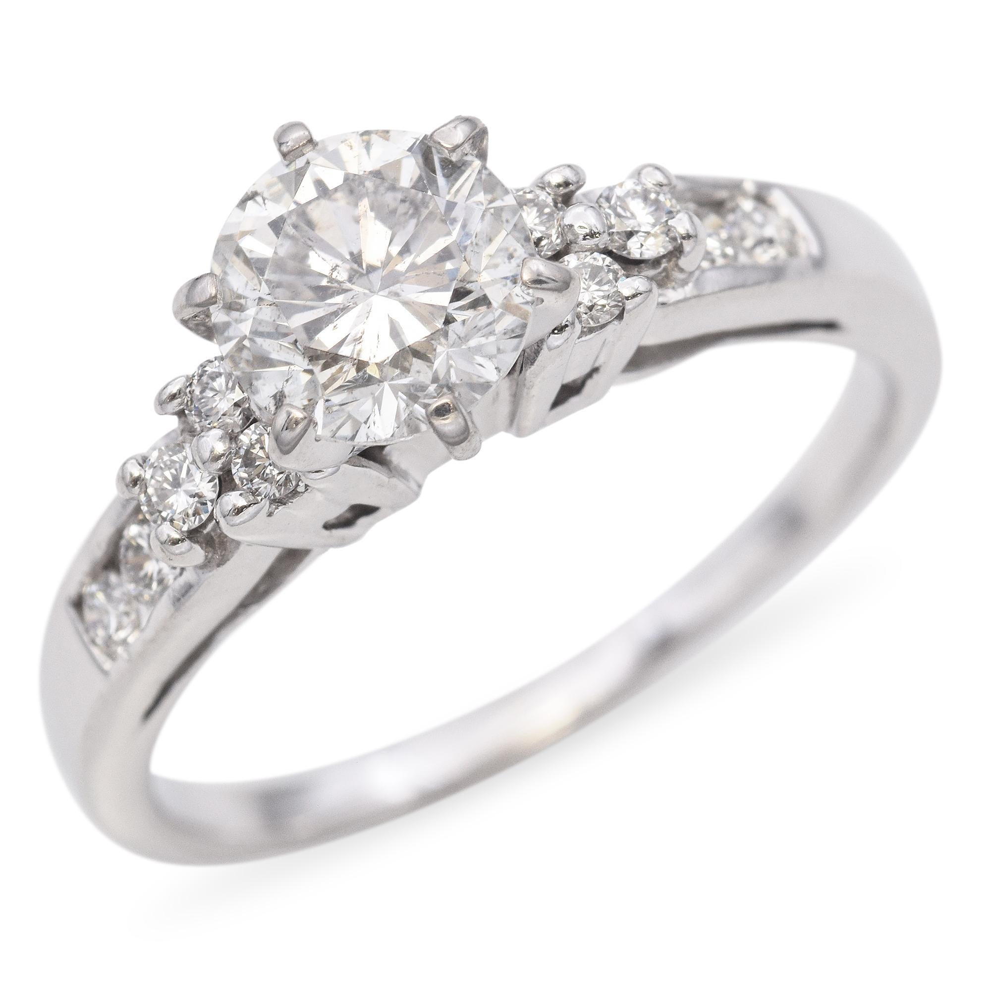 Cette bague présente un diamant brillant de taille ronde en son centre, flanqué de diamants ronds plus petits de chaque côté, créant un éblouissant spectacle de lumière et d'élégance. La bande est élégante et polie, ce qui rehausse l'éclat des
