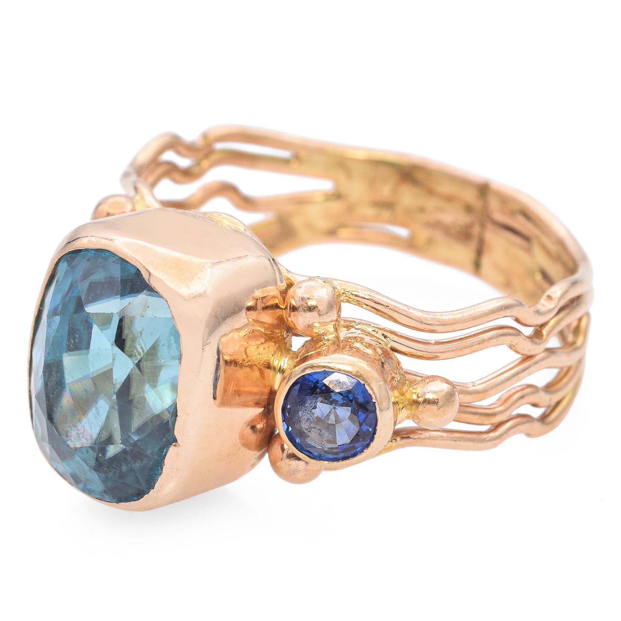 This striking ring showcases a large, vibrant teal blue topaz, expertly cut to catch the light. The central stone is flanked by a smaller, deep blue sapphire, adding depth and contrast. The setting features a unique, multi-band design in warm gold