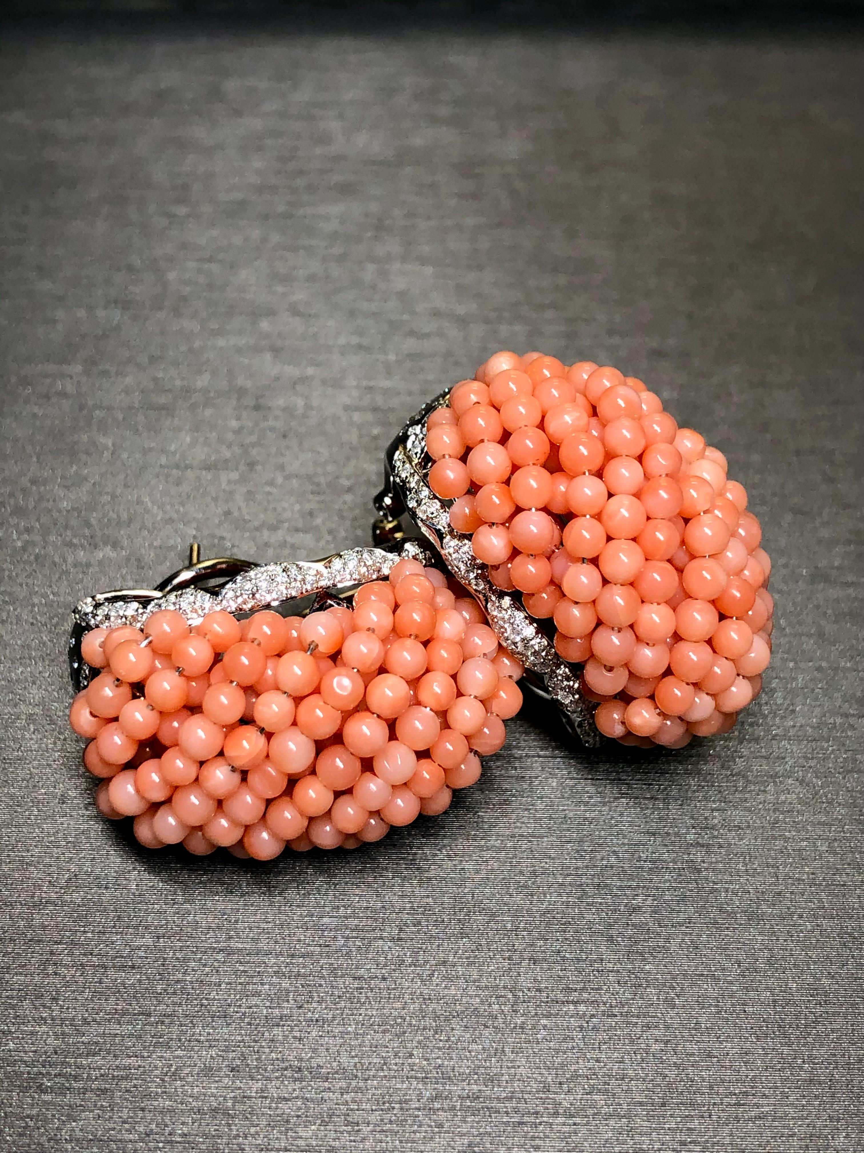 An impressive and bold pair of large huggies done in 18K white gold and set with approximately 2cttw in H-I color Si1-2 clarity round diamonds as well as numerous natural pink coral spheres intricately wired together throughout the center portion.