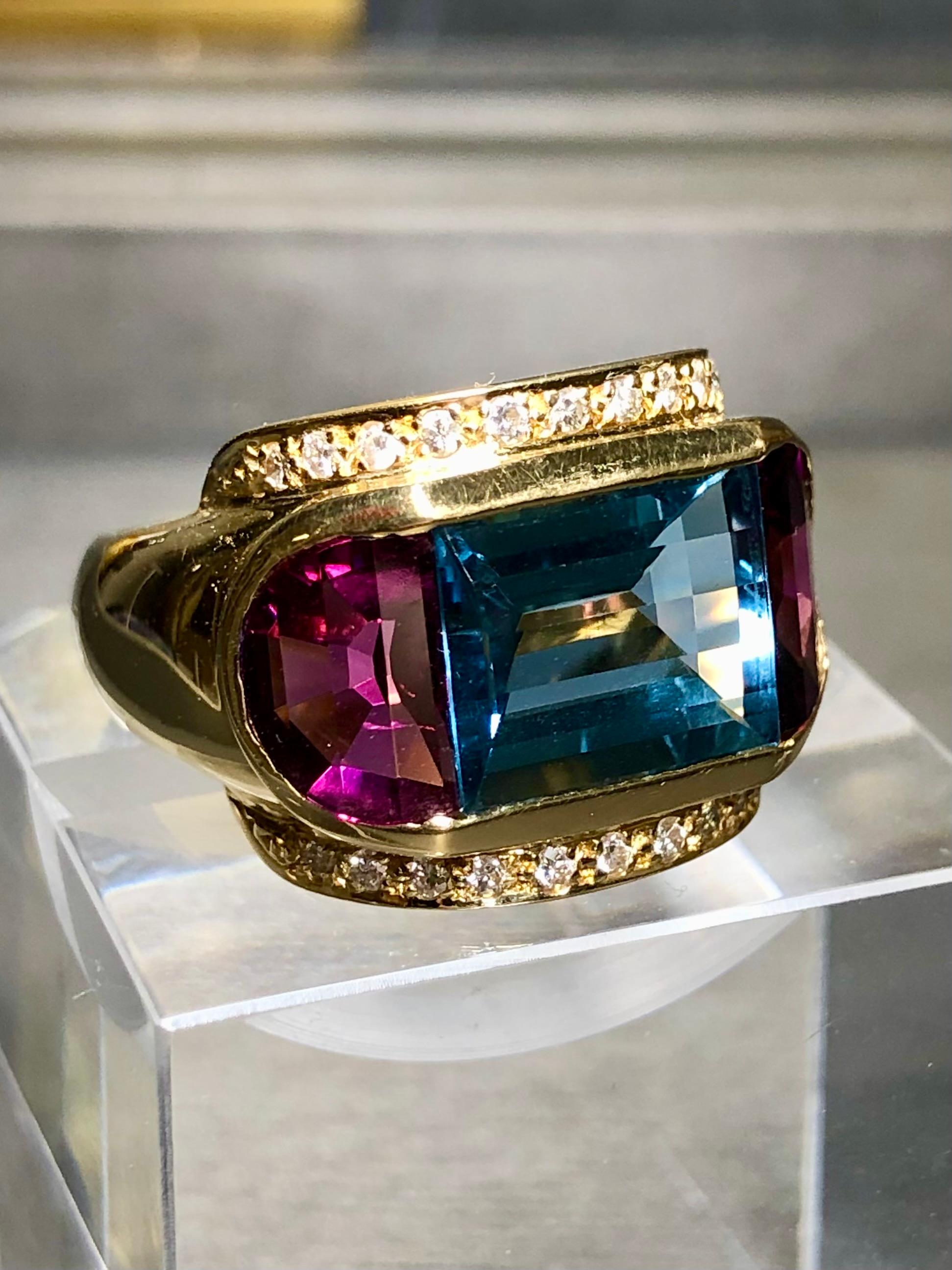 A cocktail ring that just pops! Done in 18K yellow gold, this bold ring boasts a 7ct deep blue topaz flanked by two large half-moon rhodolite garnets weighing approximately 8cttw with approximately .22cttw in H-I color Vs1-Si1 clarity diamonds on