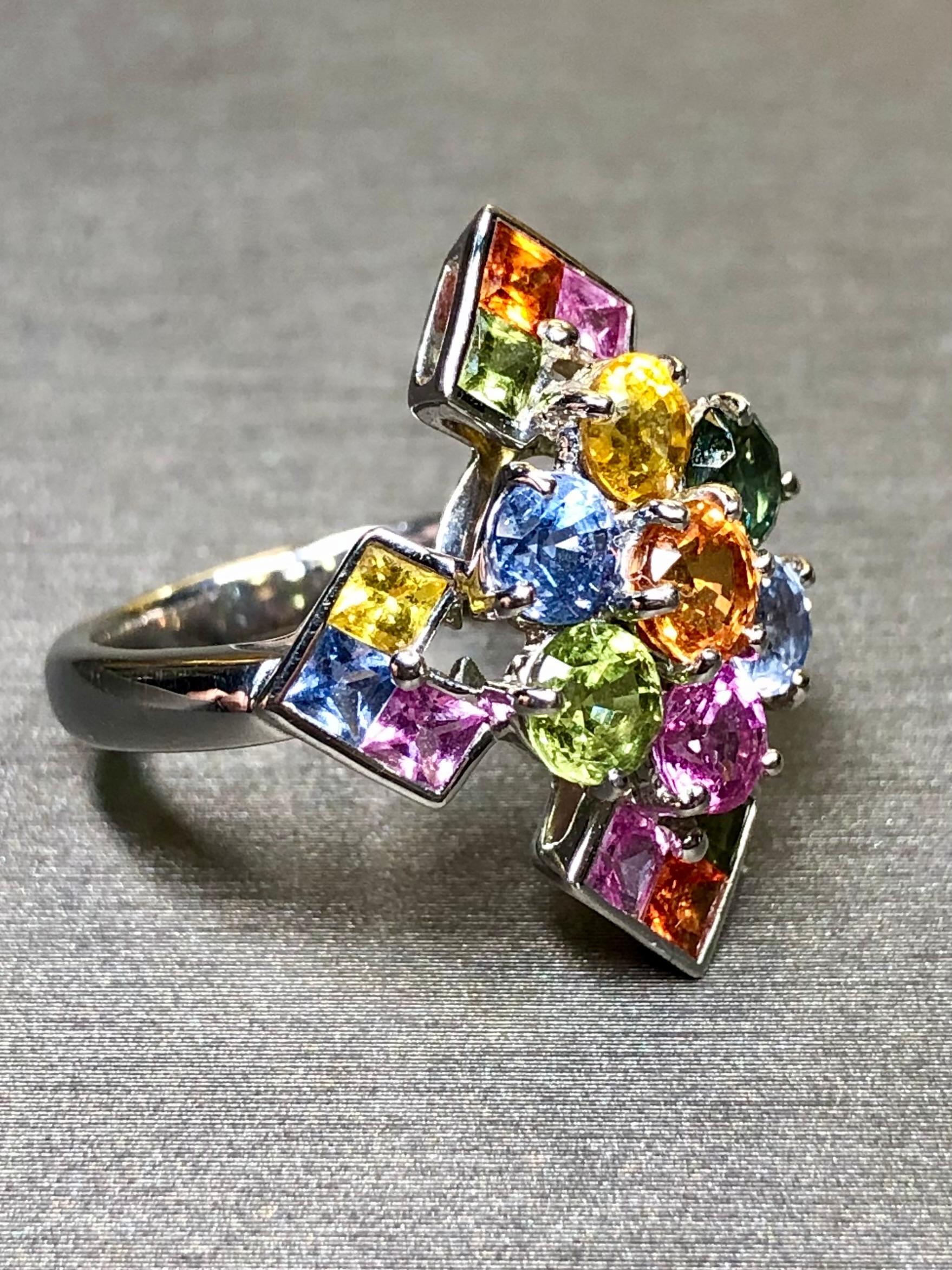 A bold and colorful contemporary ring crafted in 18K white gold and set corner to corner with 5.05cttw (stamped) in multicolored natural sapphires (heat only). It is set with both square cut as well as round cut sapphires in both a shared prong and