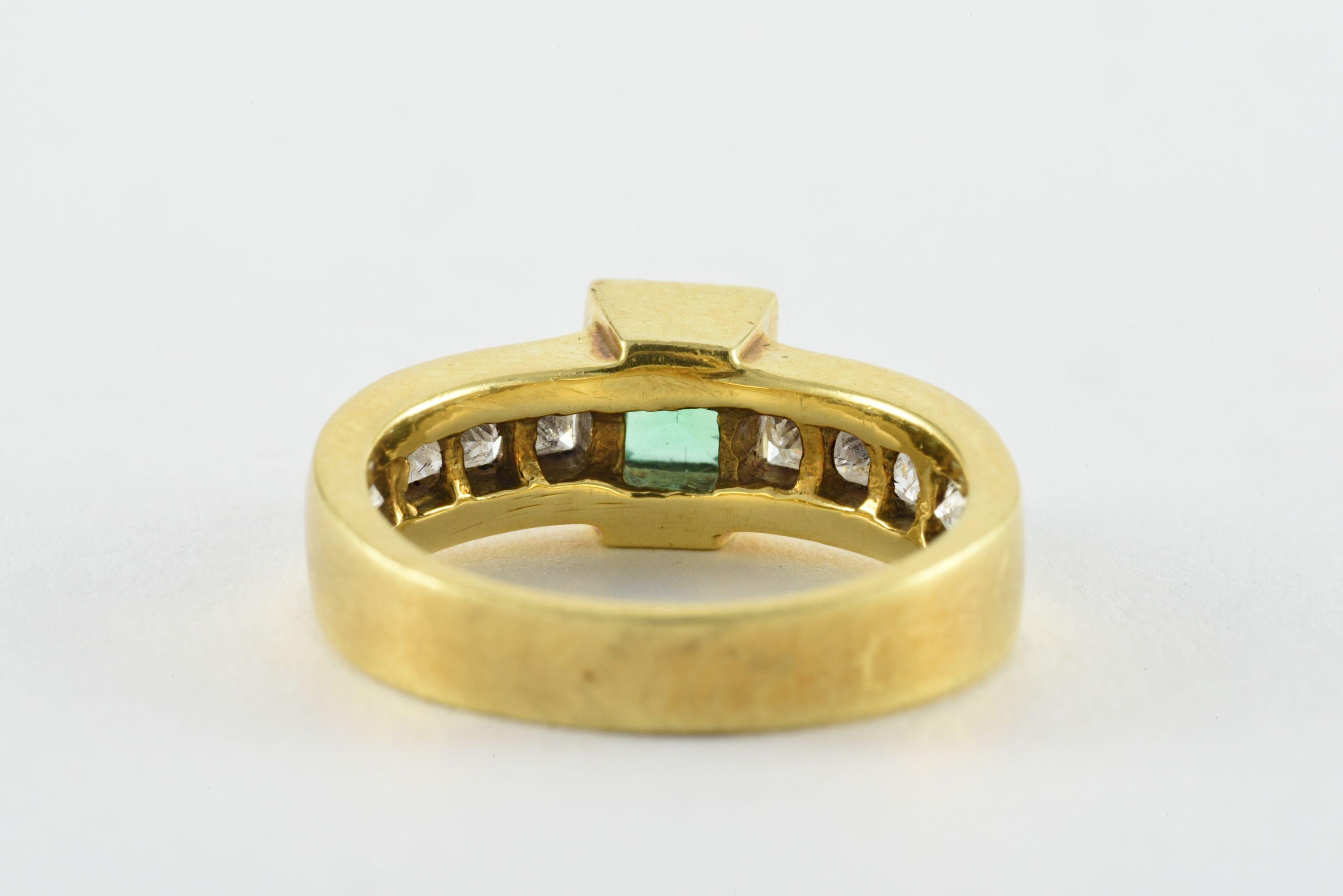 A vibrant green emerald-shaped Colombian emerald measuring approximately 0.50 carats is the centerpiece of this estate band set in a substantial 18kt yellow gold mounting, accented on the shoulders with ten princess cut diamonds, in a channel