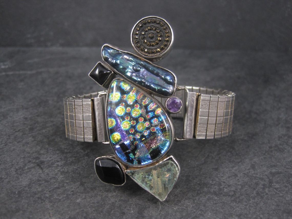 Cet énorme et magnifique bracelet de créateur est composé d'une pièce centrale en argent sterling avec un magnifique verre d'art, une perle de paon biwa, de la tourmaline, de l'onyx et de l'améthyste.
Il s'agit d'une création de Patti Quinn, artiste