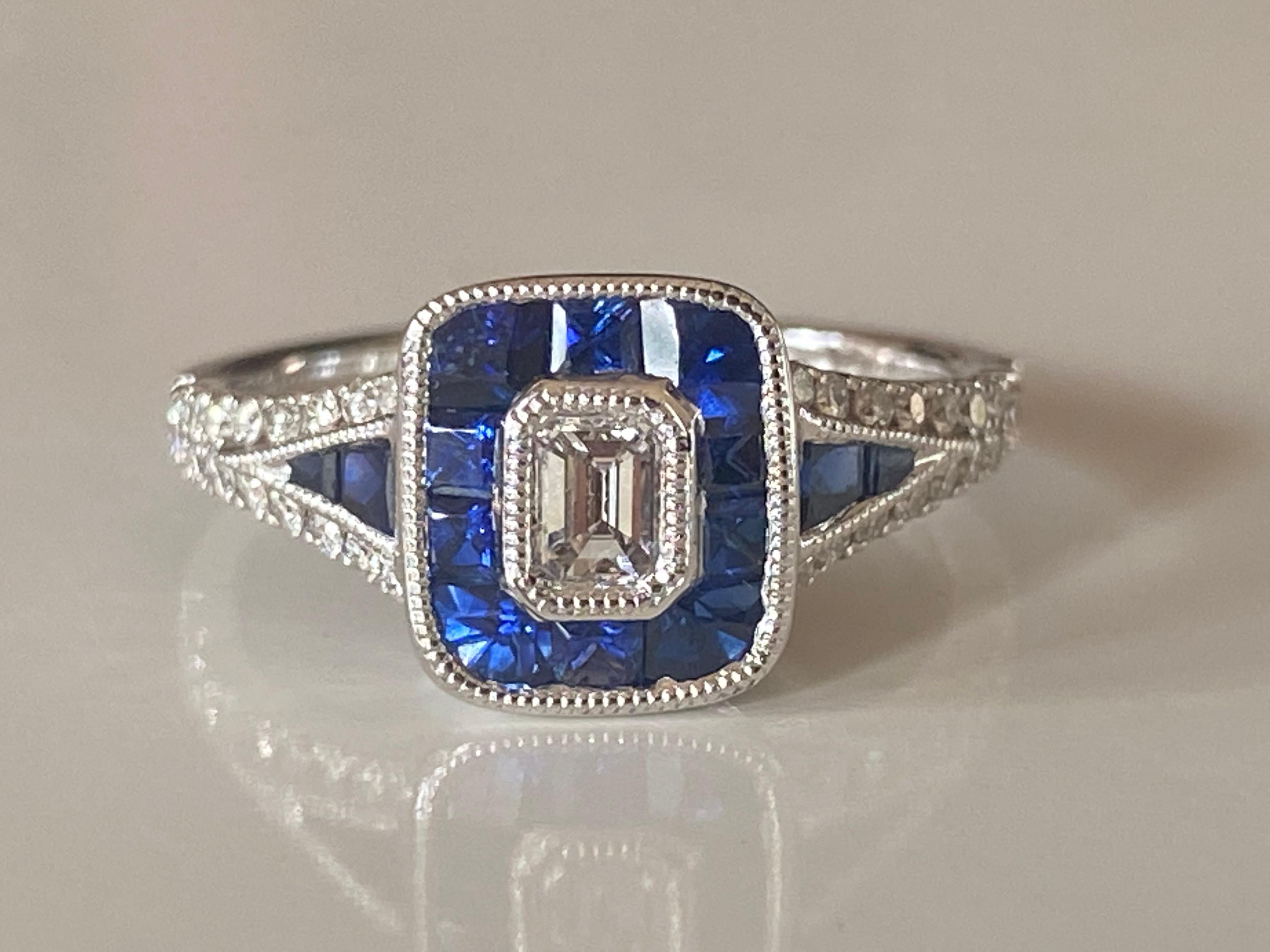 This sparkling gem set in 18kt white gold is designed around an emerald cut white diamond center stone surrounded by a halo of calibrated blue sapphires. Additional sapphires adorn the shoulders in a tapered triangular design lined by glistening