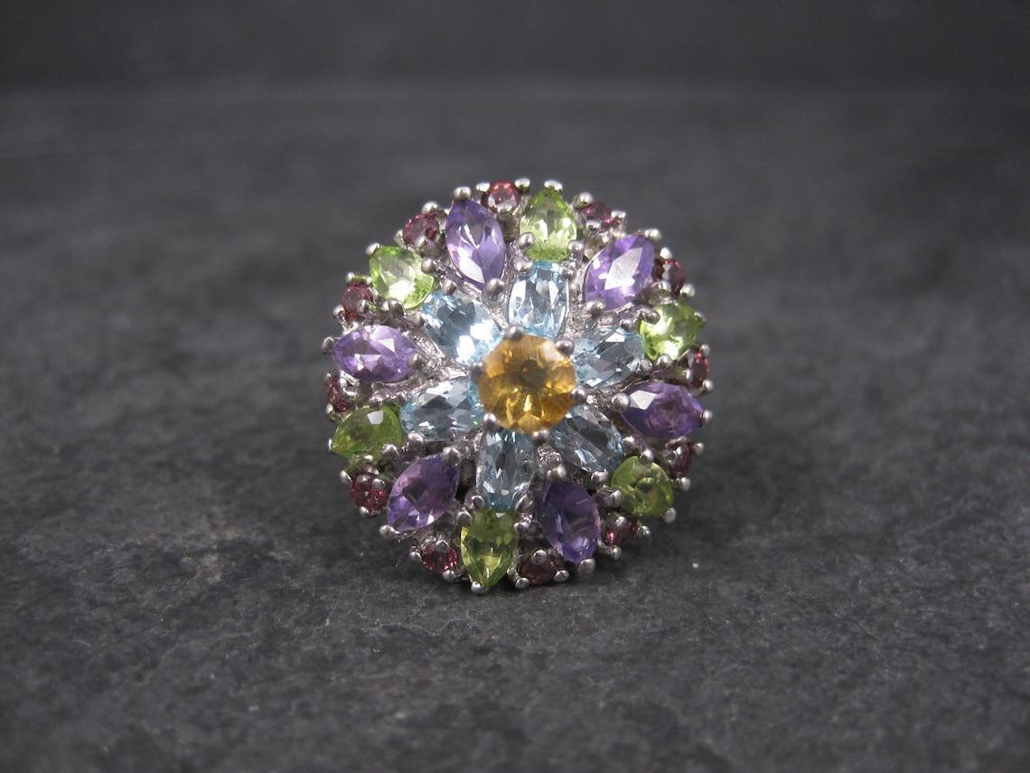 This gorgeous ring is comprised of sterling silver.
It features a combination of a 6mm medium yellow, round cut citrine, 6 oval 3x5mm blue topaz, 6 marquise cut 3x6mm amethysts, 6 pear cut 3x5mm peridots, and finally, 12 round 2mm raspberry