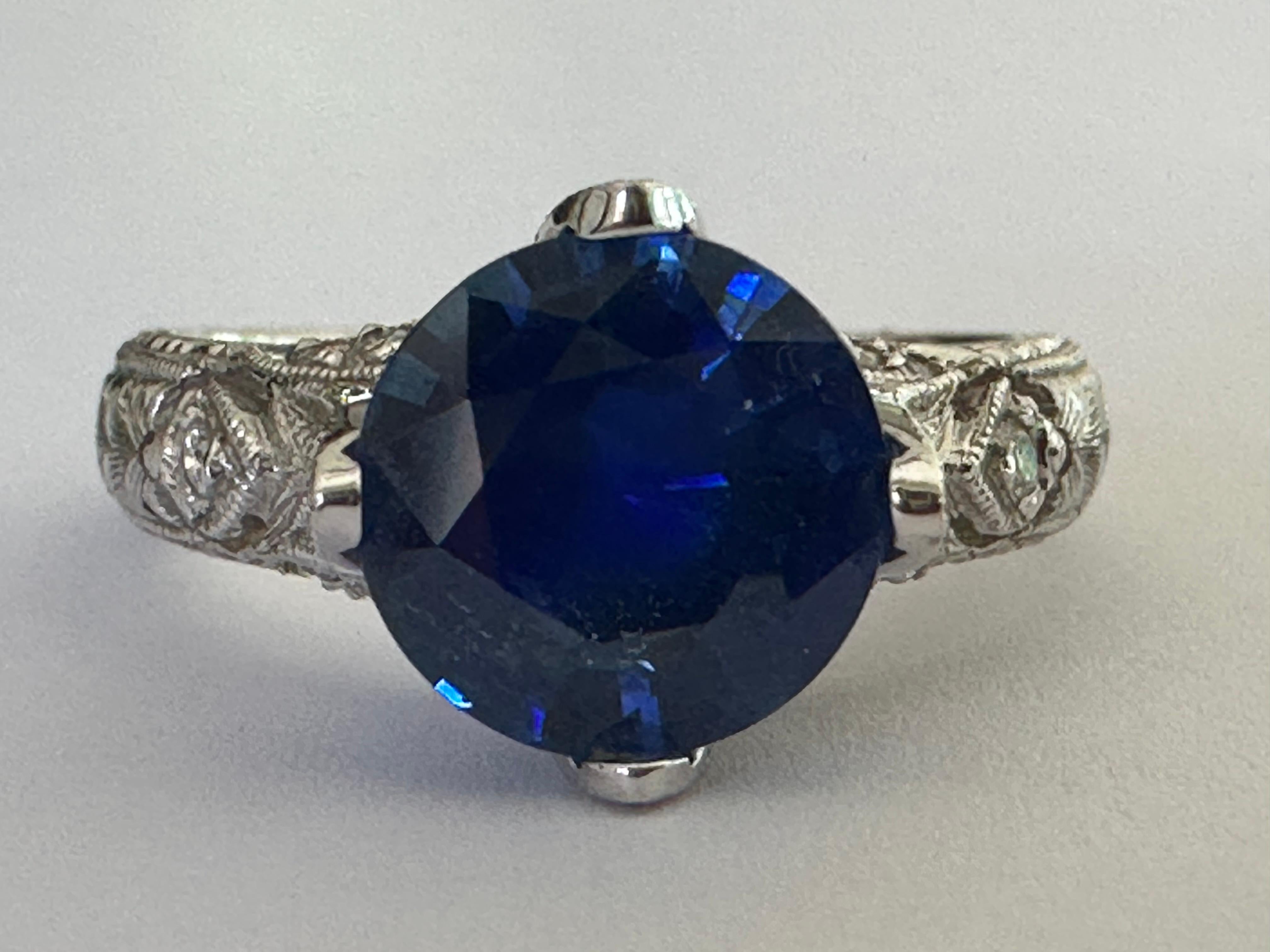 This ring features a 2.45-carat natural deep blue round-shaped sapphire adorned with 18 round diamonds totaling approximately 0.23 carats. Stamped VIACHI and set in 18K white gold with delicate hand engraving along the band. 
