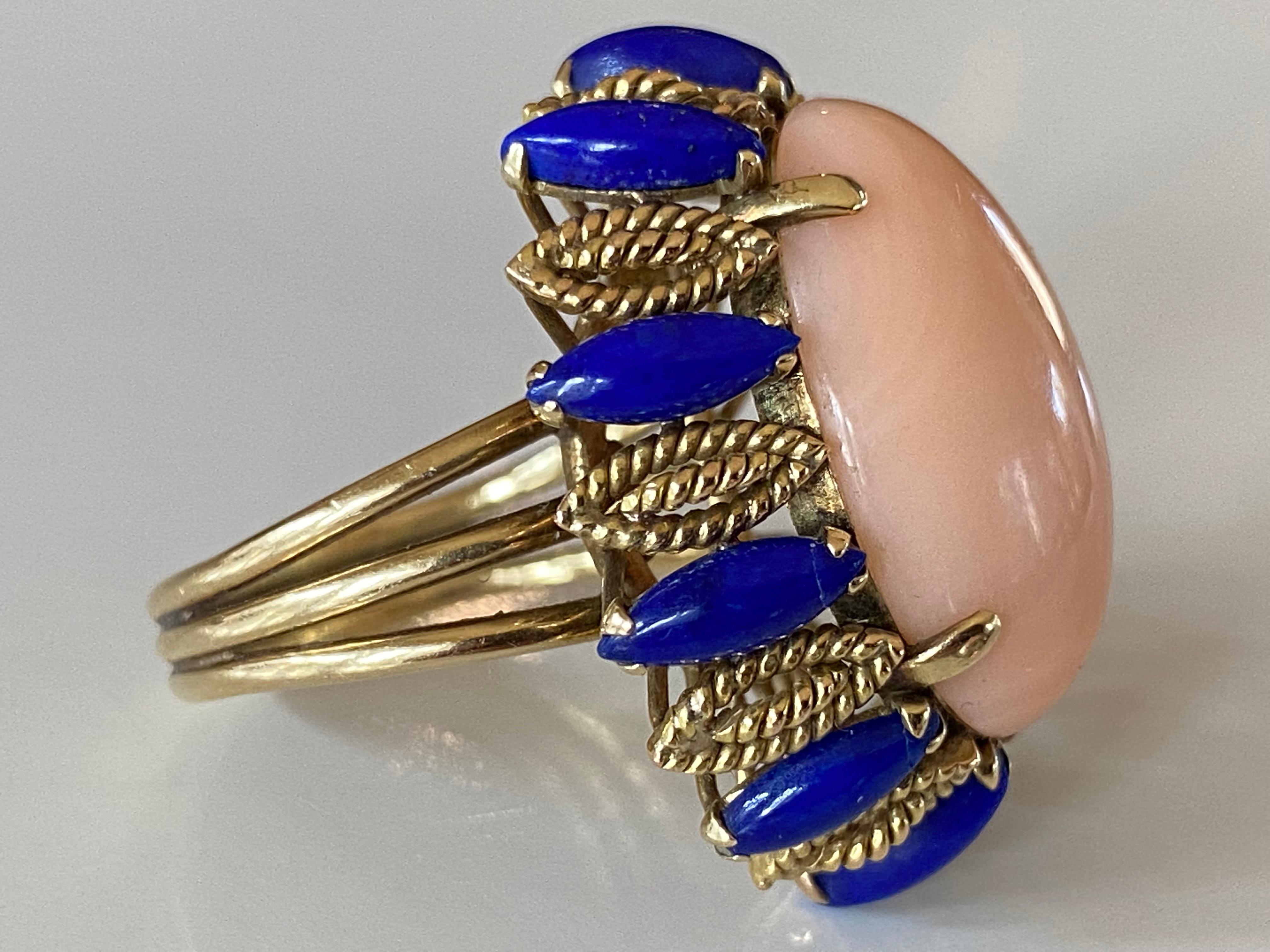 Crafted in the 1950s in 14kt yellow gold, this stunning mid-century cocktail ring features a natural angel skin coral oval-shaped cabochon measuring approximately 14 x 20mm encircled by ten marquis-shaped lapis lazuli stones. Matching earrings