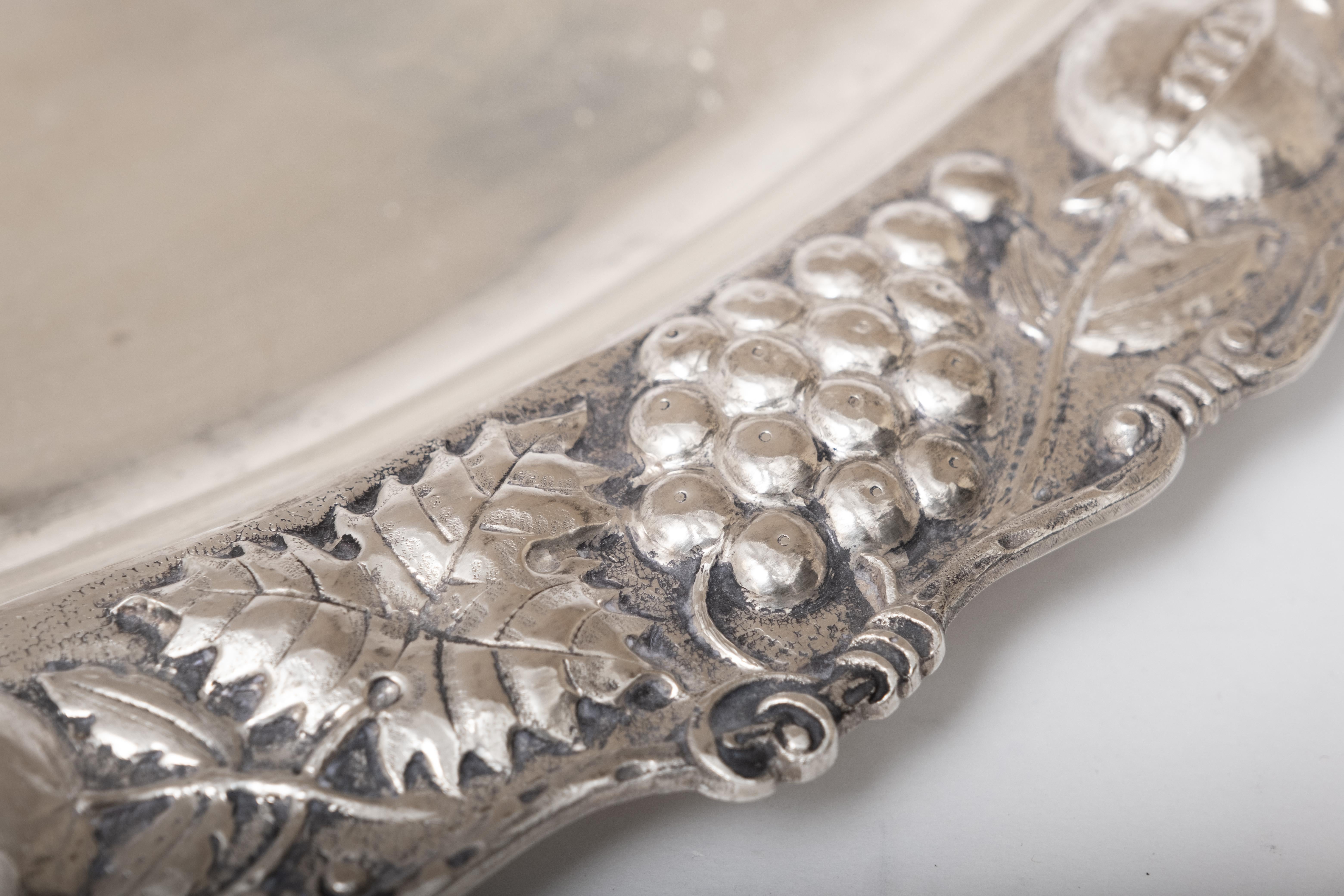 Presenting a beautifully crafted 900 grade silver platter, showcasing a captivating repoussé design along its edges. This artistic motif features an array of intricate flowers framed by a curvilinear grape vine pattern, adding an elegant touch to