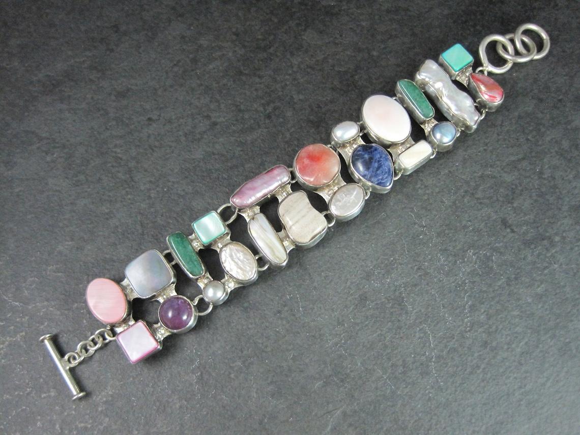 This gorgeous estate bracelet is sterling silver.
It features a combination of stones including mother of pearl in various colors, pearls, jade, blister pearls, biwa pearls, sodalite and more.

Measurements: 1 1/8 inches wide - Can be toggled at 7,