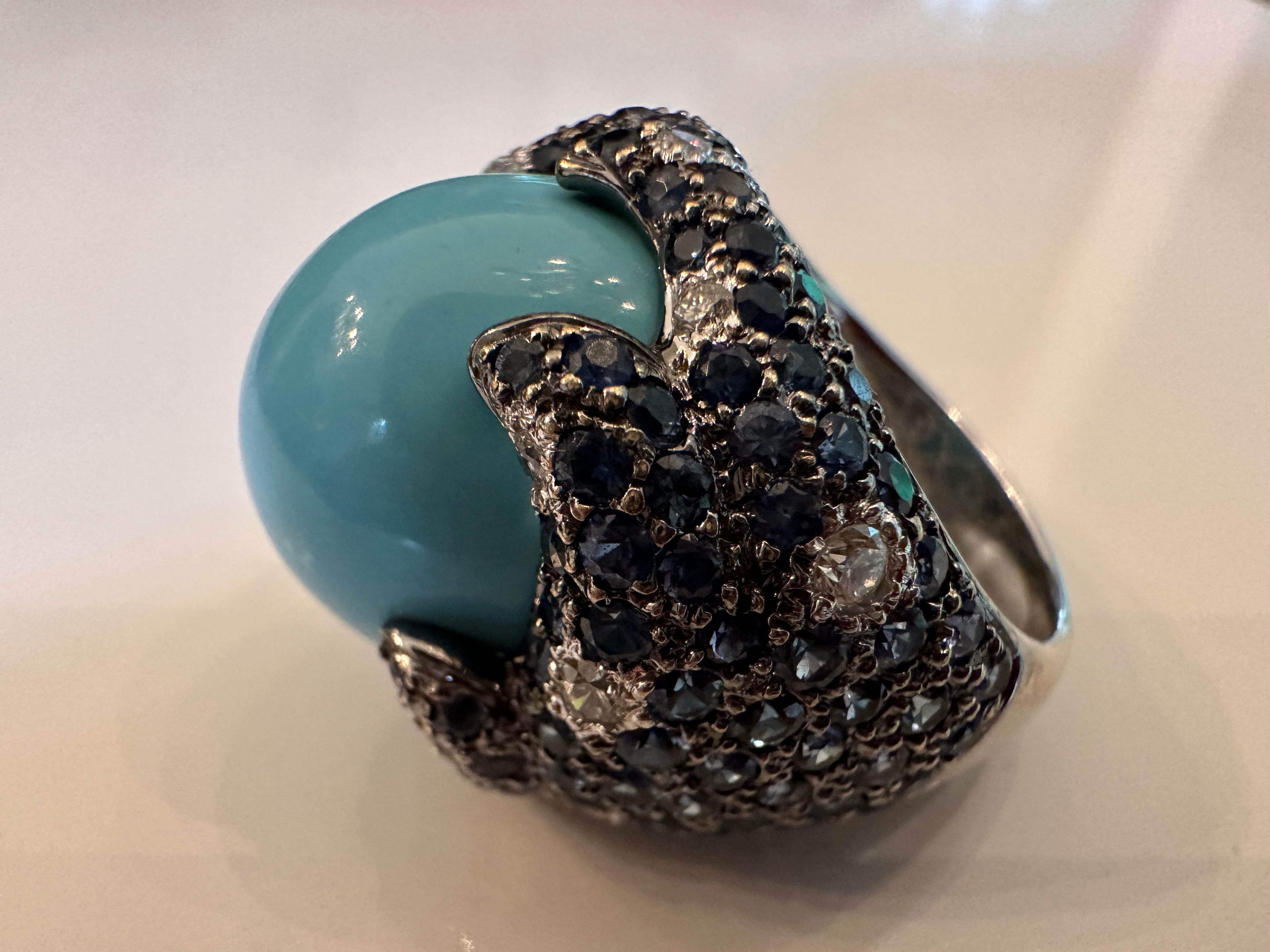 This striking cocktail ring features a natural turquoise center stone measuring approximately 16.5x18.7mm surrounded by round-shaped blue sapphires and round diamonds artfully arranged in a unique dome shape. Stamped 18K, 750. Set in 18K white gold.