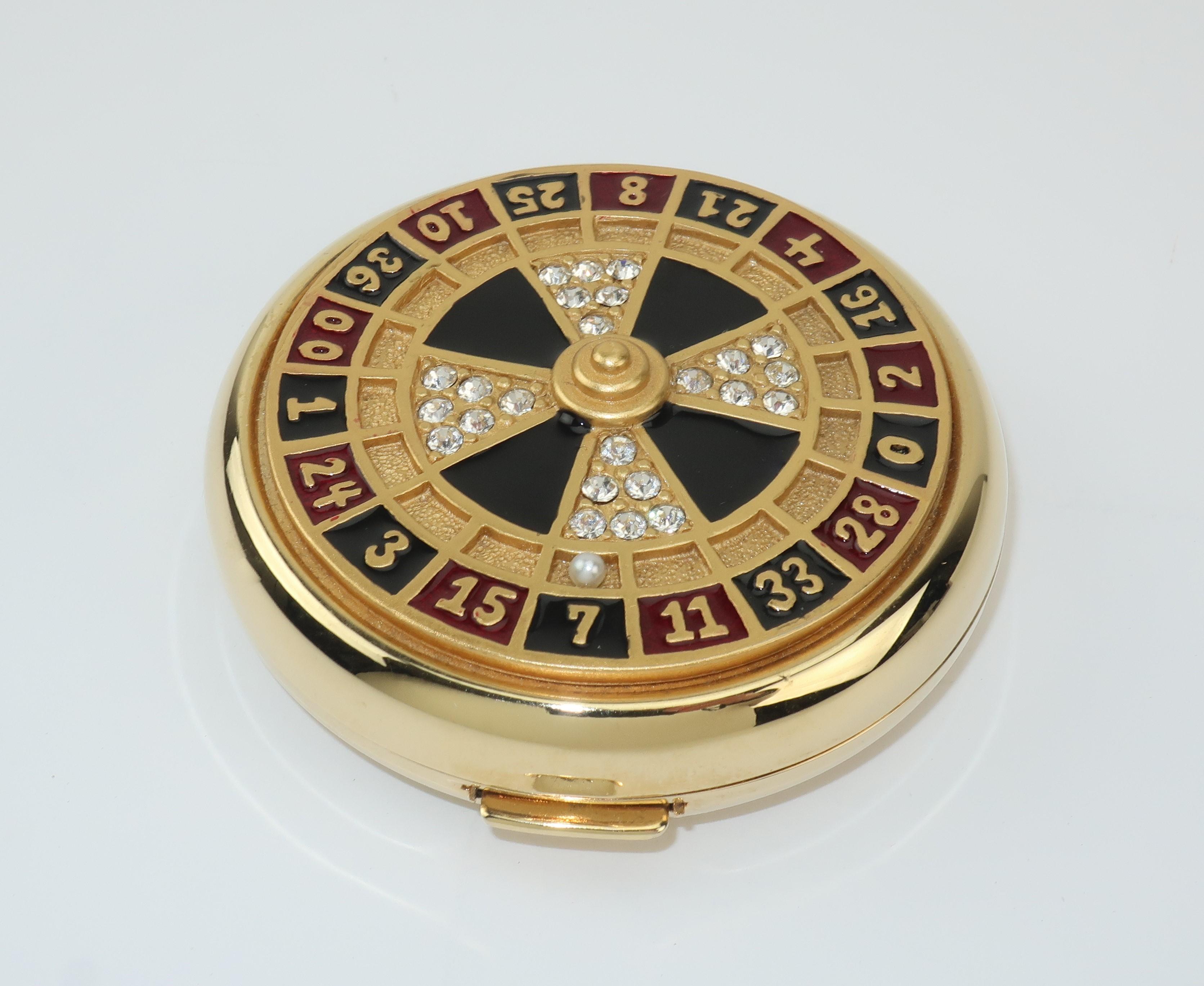 Lady luck! A collectible powder compact from Estee Lauder in the shape of a roulette wheel with black and dark red enameling accented by crystal rhinestones and a single pearl all set in a jewelry quality gold tone metal. The push button closure