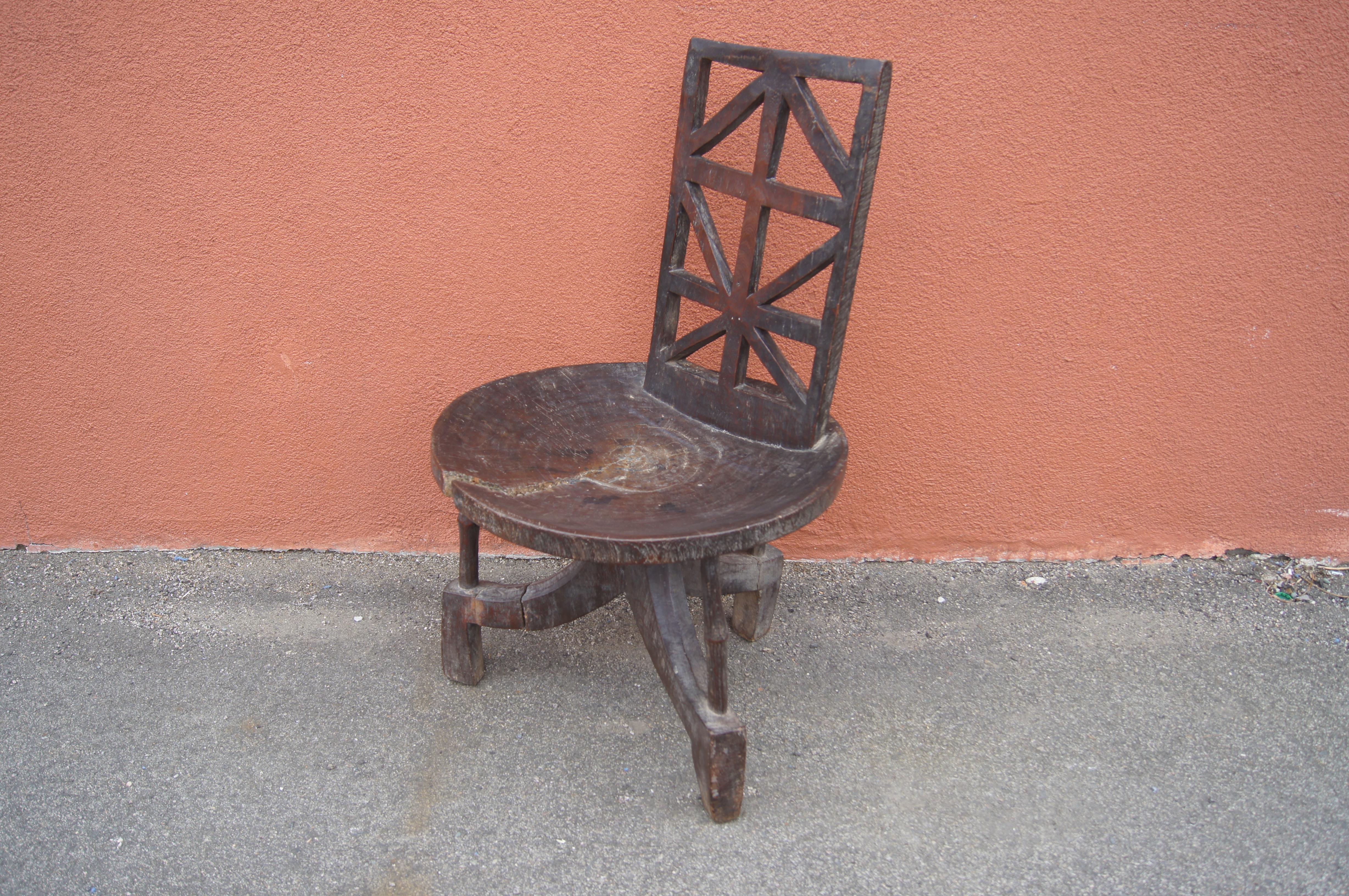 This carved wooden chair most likely comes from the Jimma people of Southern Ethiopia. A capacious slightly concave seat sits on tripod base connected by slender rods. The high rectangular seatback has a geometric openwork design.