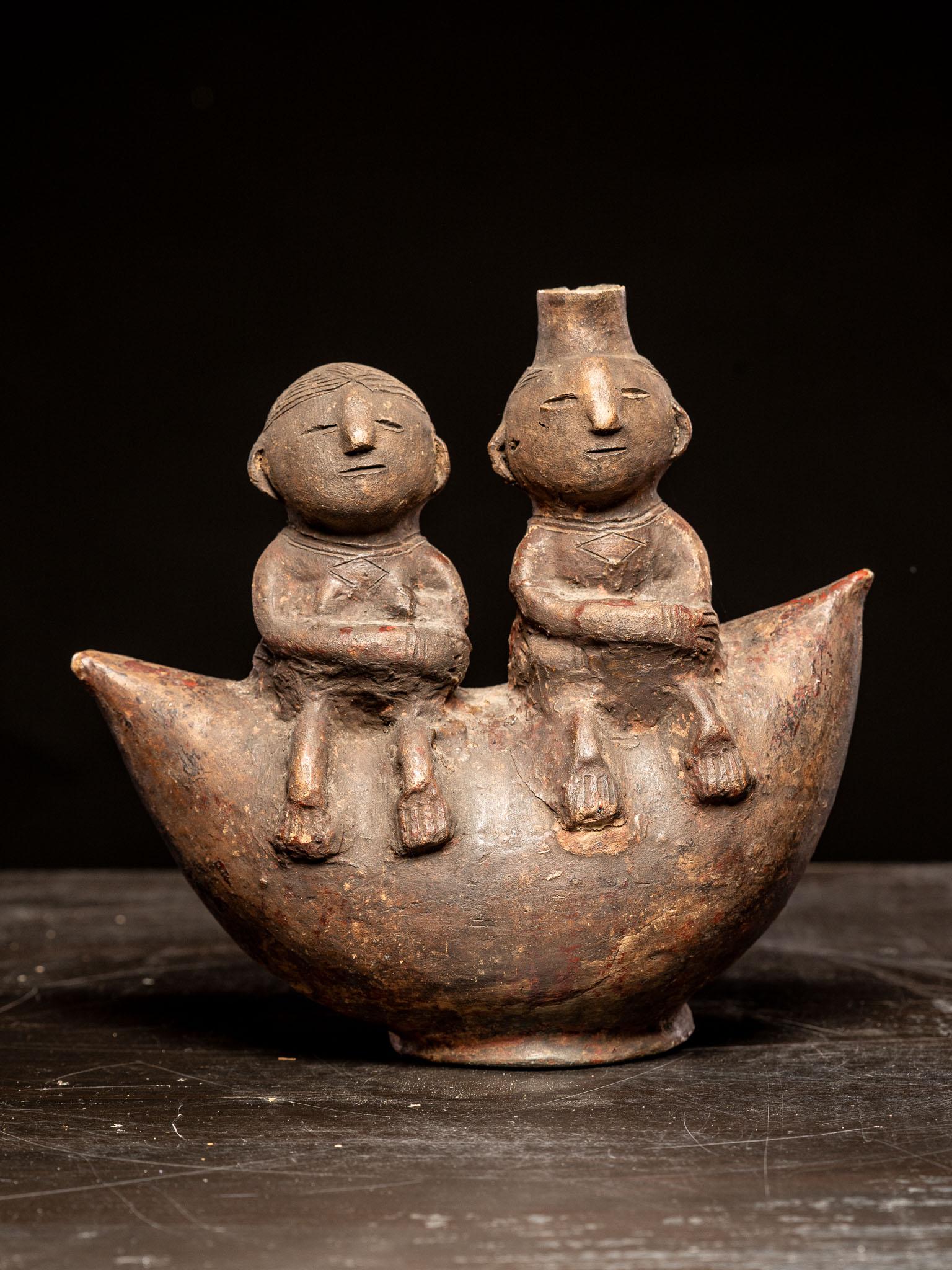 Terracotta pot of roughly crescent shape featuring a pair of naively modelled human figures seated on the concave edge, each with arms folded to the abdomen and feet pointing downwards, the female with a flat incised coiffure, the male with a short