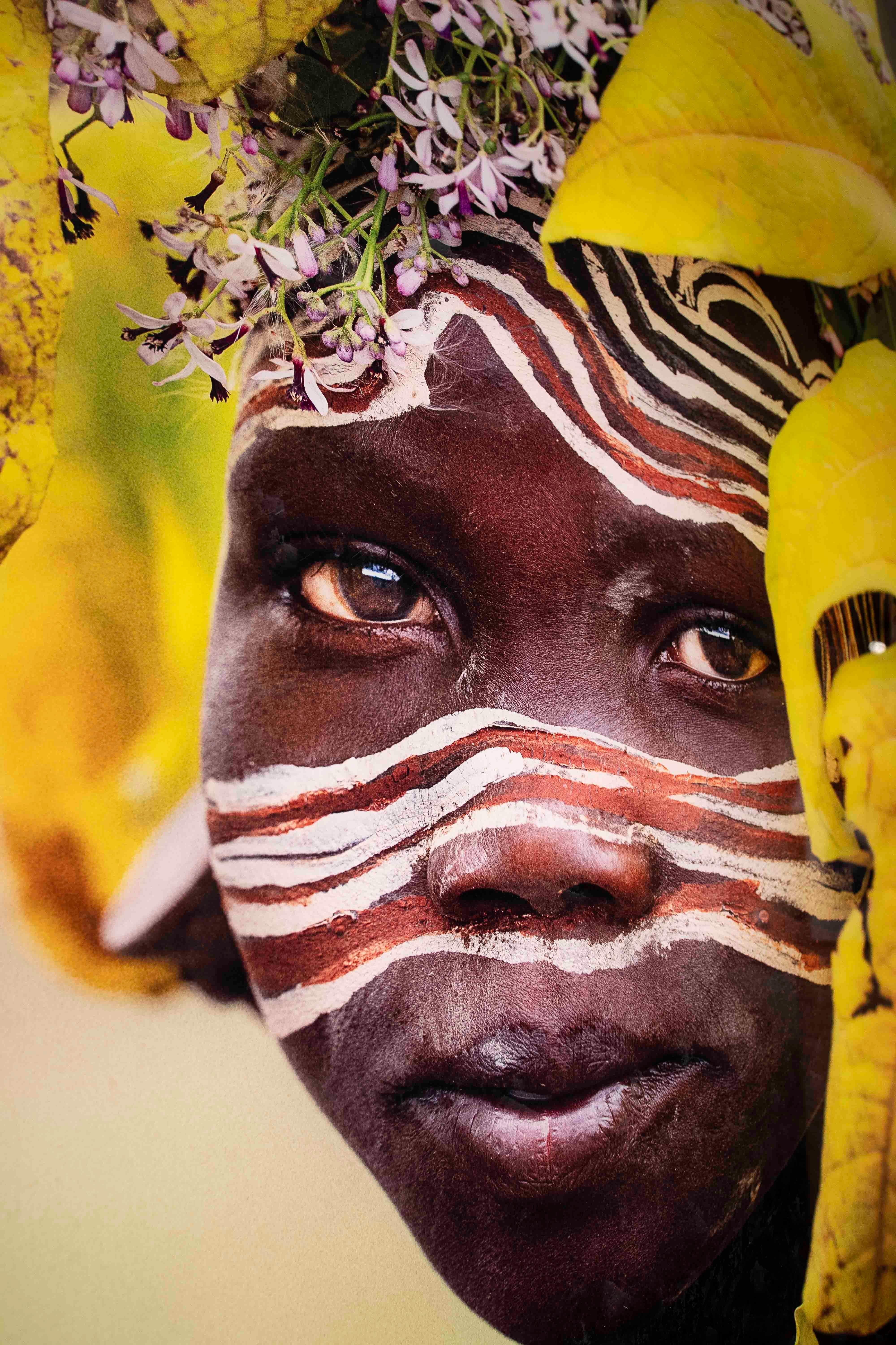 Ein atemberaubendes Foto, das die Essenz der Kultur und die Verbindung zur Natur einfängt. Das Porträt zeigt eine Frau, die mit gelben Blättern und fliederfarbenen Blüten geschmückt ist, ergänzt durch rote und weiße Gesichtsbemalung, die eine tiefe