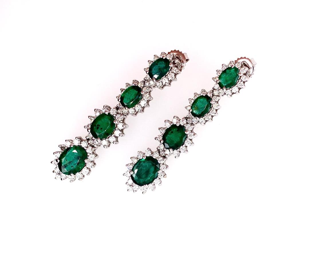 Estos elegantes pendientes presentan un verde intenso,   Esmeraldas verdes talla oval rodeadas de un halo de diamantes redondos talla brillante engastados en oro blanco de 18 quilates. Cada pendiente consta de cuatro racimos graduados de esmeraldas