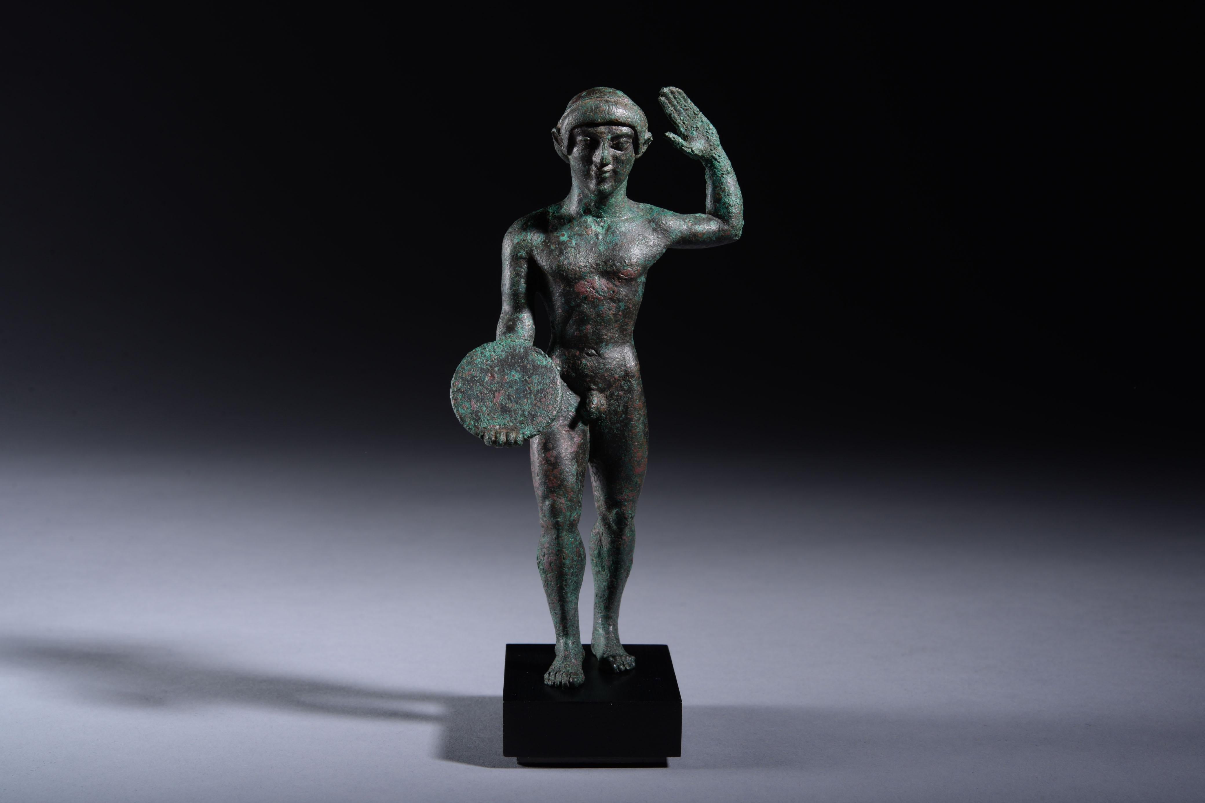 A remarkably fine example of Archaic sculpture.  A figure of a discophoros, or discus bearer, standing with his right leg forward, gripping a discus in his right hand, the left arm raised with an open palm. Described with admiration in Münzen und