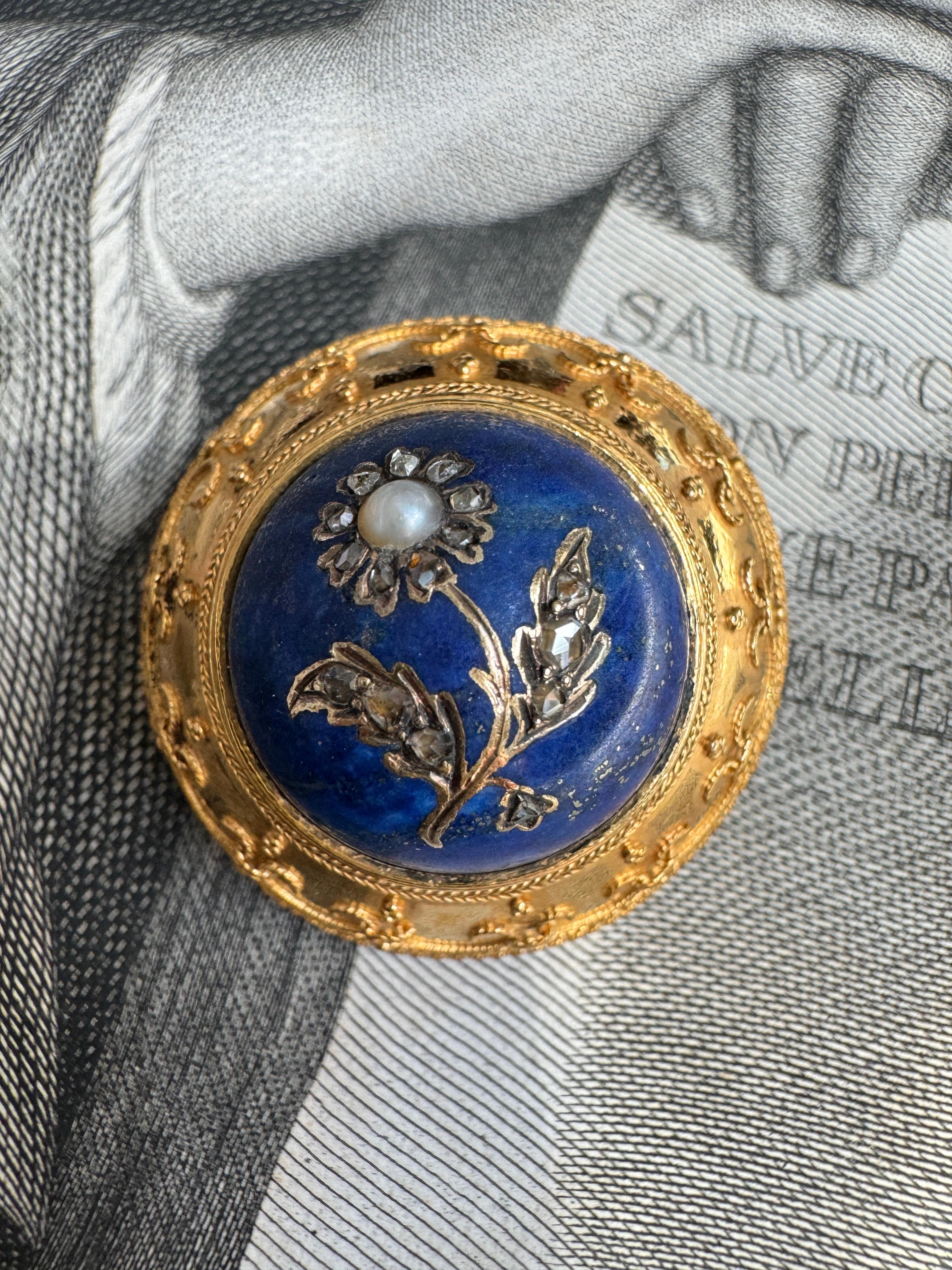 This delightful Etruscan Revival brooch, circa 1880, features a high profile lapis lazuli cabochon inlaid with a glittering rose-cut diamond daisy. Rendered in velvety 15 karat gold, the frame is artfully adorned with delicate granulation and