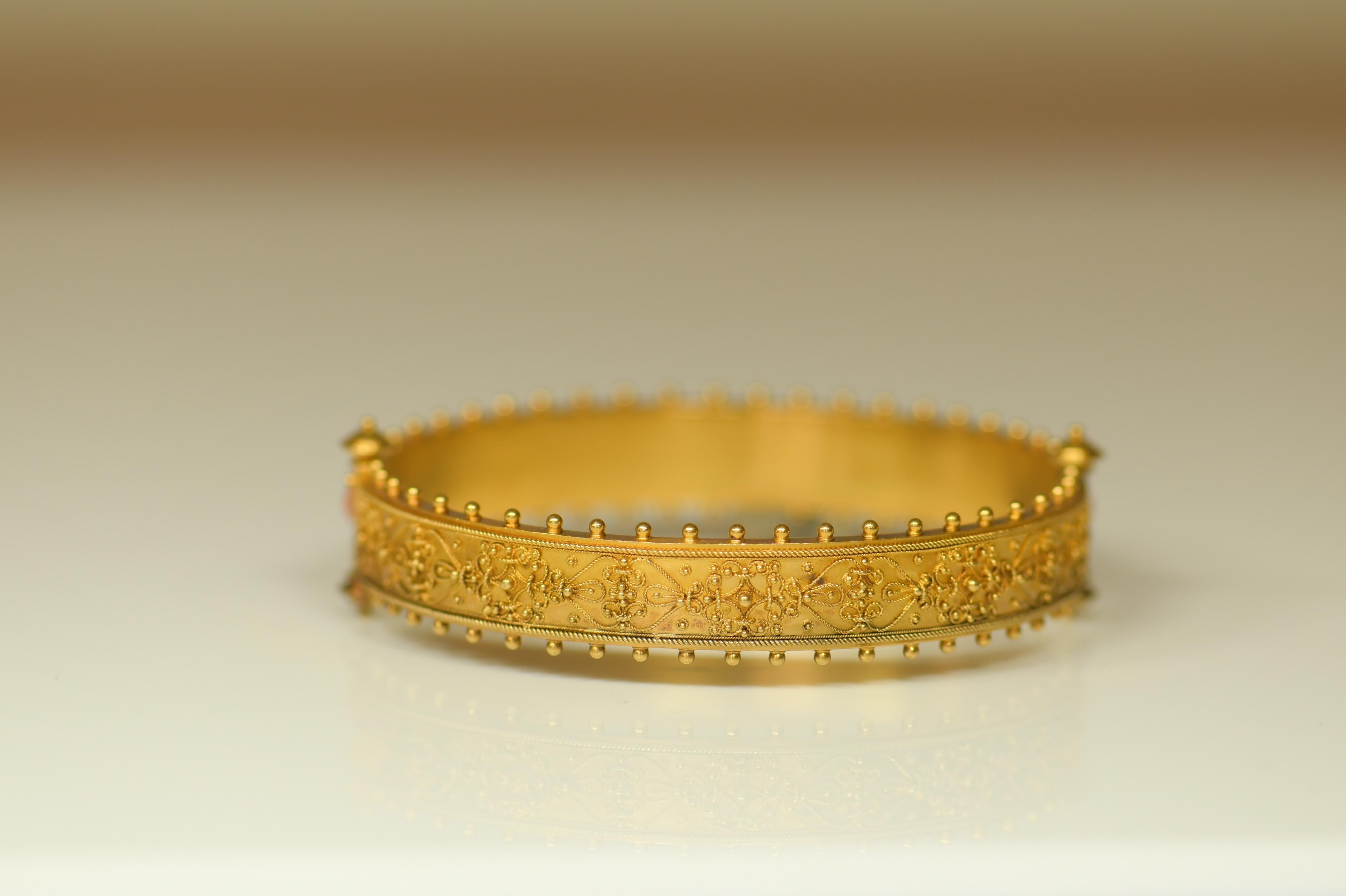 An Etruscan Revival gold and coral hinged bangle, set to the one side with eleven cabochon-cut corals, surrounded by wire-work decoration of geometric design, the other side with wirework of similar design,  all with gold bead edge, circa